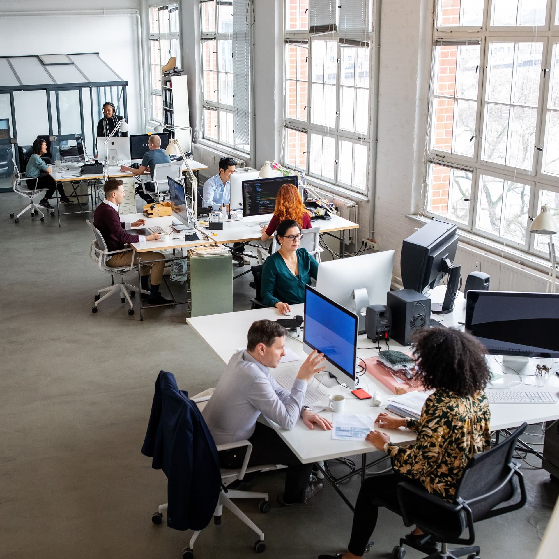 Ein modernes Großraumbüro mit Glasfront aus der Vogelperspektive. Zahlreiche Personen arbeiten am Computer an unterschiedlichen Tischen.