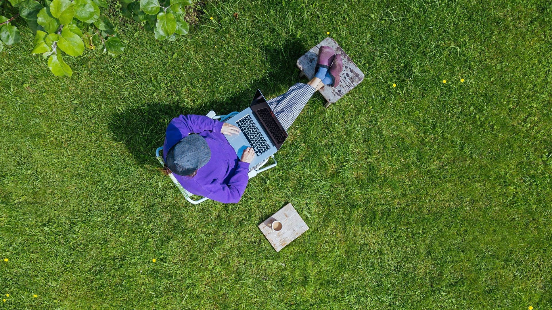 Eine junge Frau, aus Vogelperspektive fotografiert, sitzt mit ihrem Laptop auf dem Schoß und einem Kaffee neben sich bequem in ihrem Garten, auf ihrem Grundstück. 
