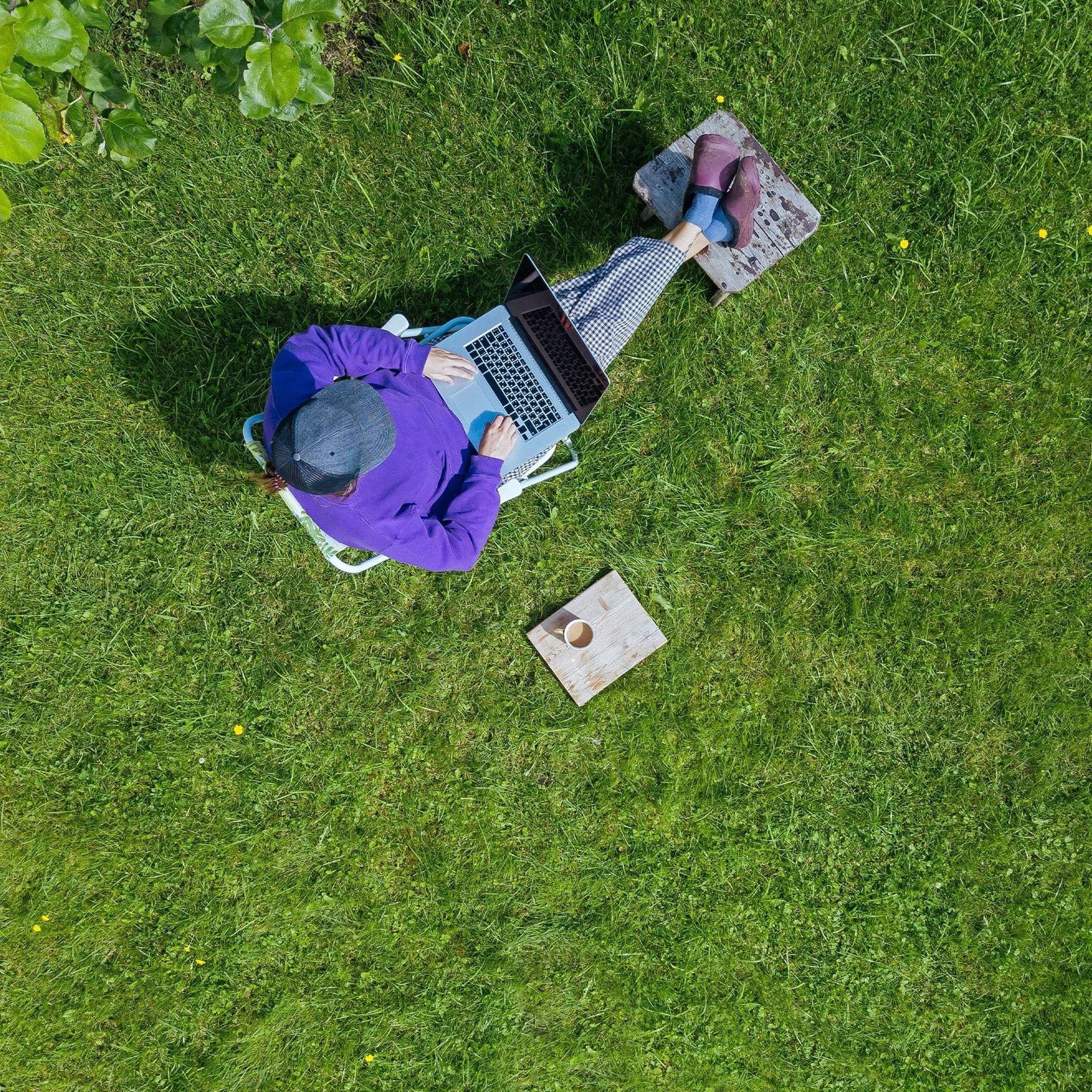 Eine junge Frau, aus Vogelperspektive fotografiert, sitzt mit ihrem Laptop auf dem Schoß und einem Kaffee neben sich bequem in ihrem Garten, auf ihrem Grundstück. 