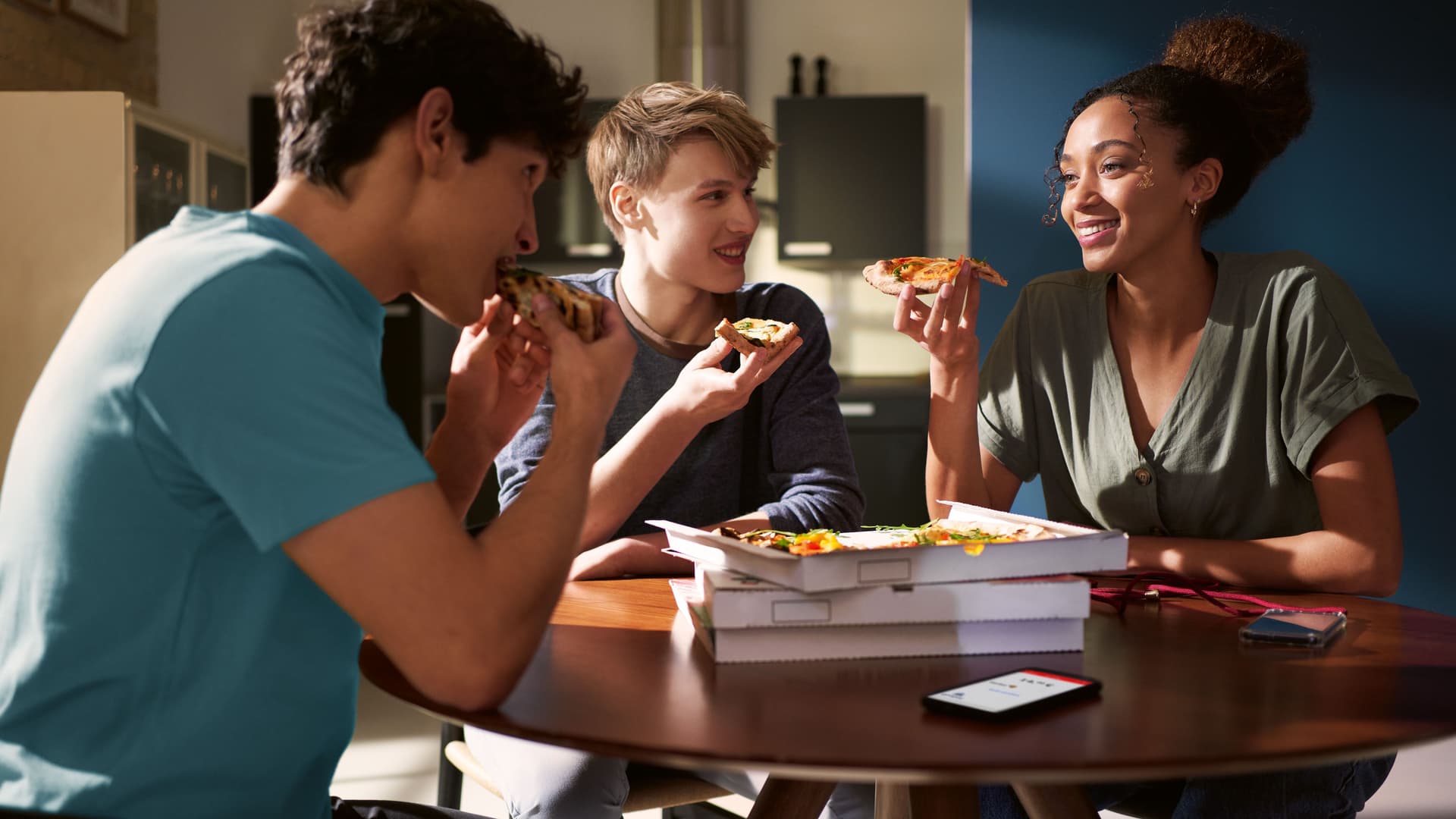 Zwei Männer und eine Frau sitzen an einem Tisch und haben Pizzastücke in der Hand. Auf dem Tisch stehen mehrere Pizzakartons.
