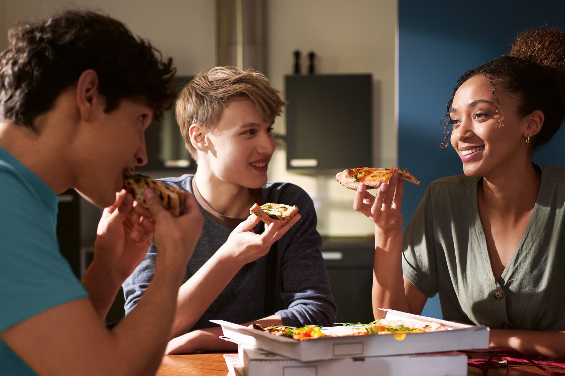 Zwei Männer und eine Frau sitzen an einem Tisch und haben Pizzastücke in der Hand. Auf dem Tisch stehen mehrere Pizzakartons.