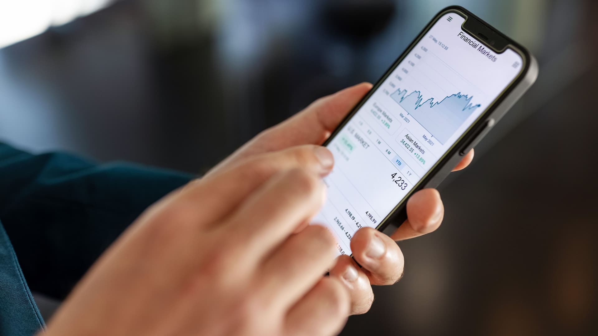 Nahaufnahme männlicher Hände mit einem Smartphone, das Aktienkurse zeigt.