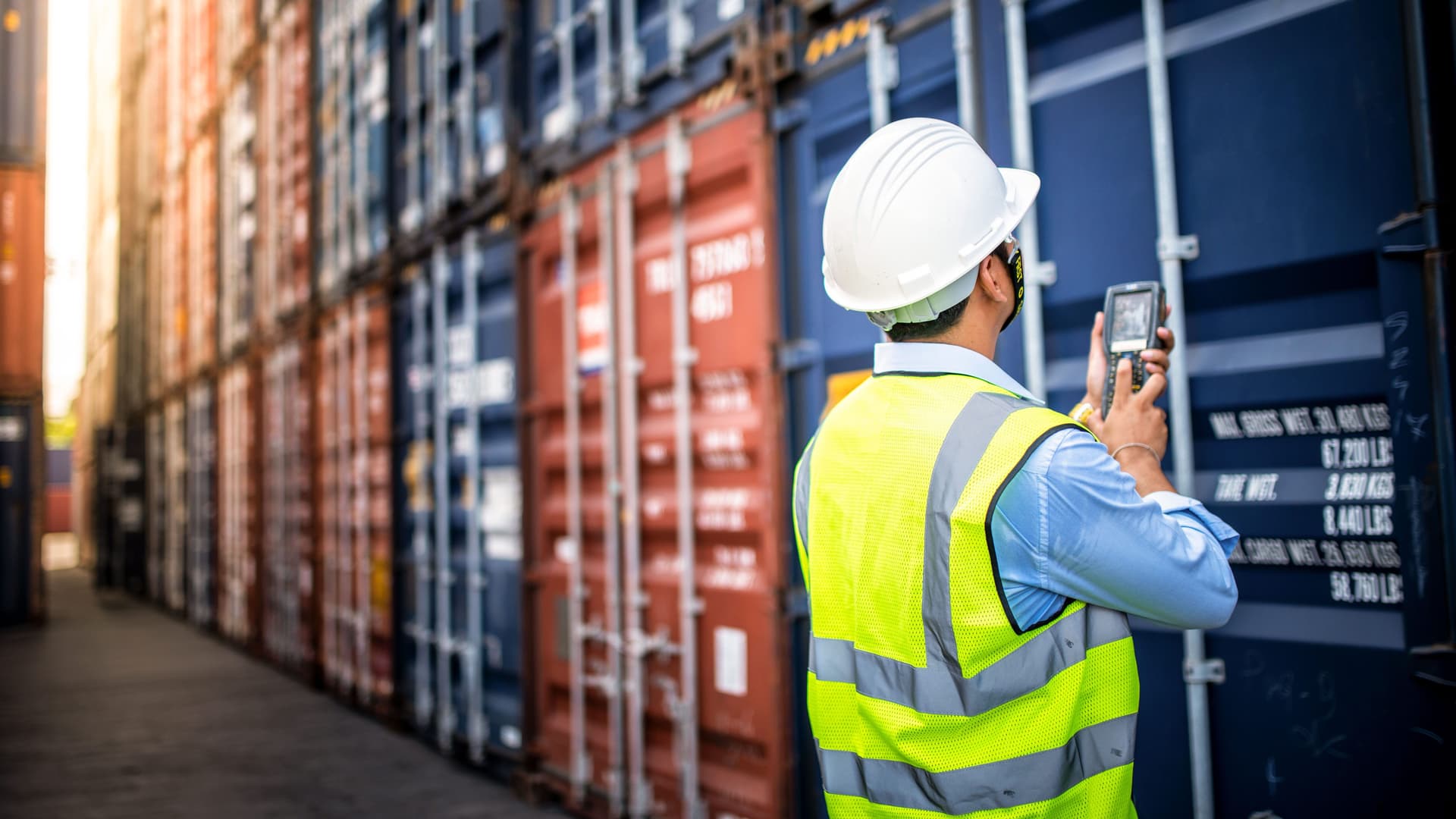 Ein Logistiker mit Helm, Warnweste und Scanner steht mit dem Rücken zur Kamera vor übereinandergestapelten Containern.