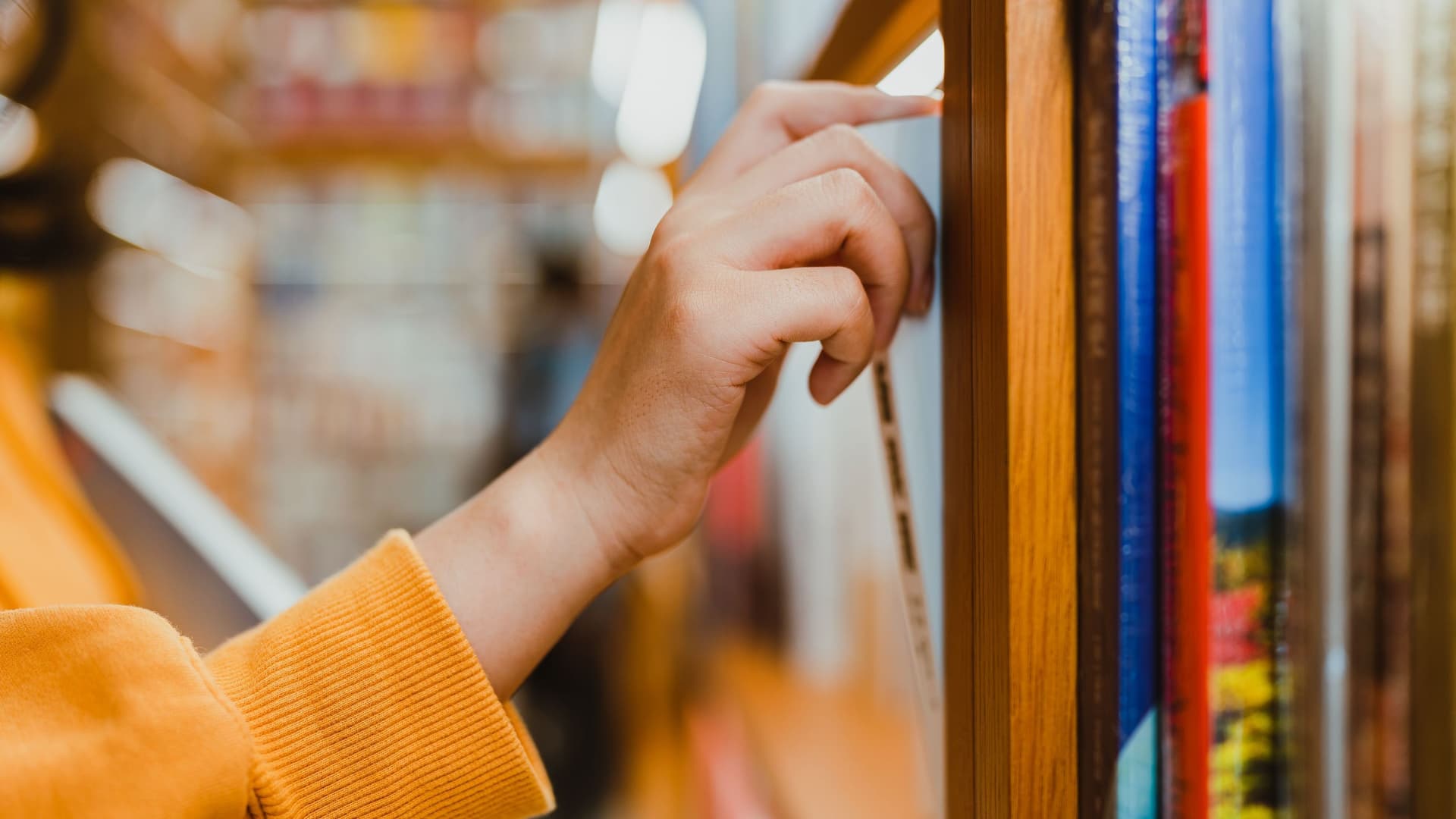 Seitliche Nahaufnahme von einem Arm mit orange farbenen Pullover. Die Hand zieht ein Buch aus einem Bücherregal.