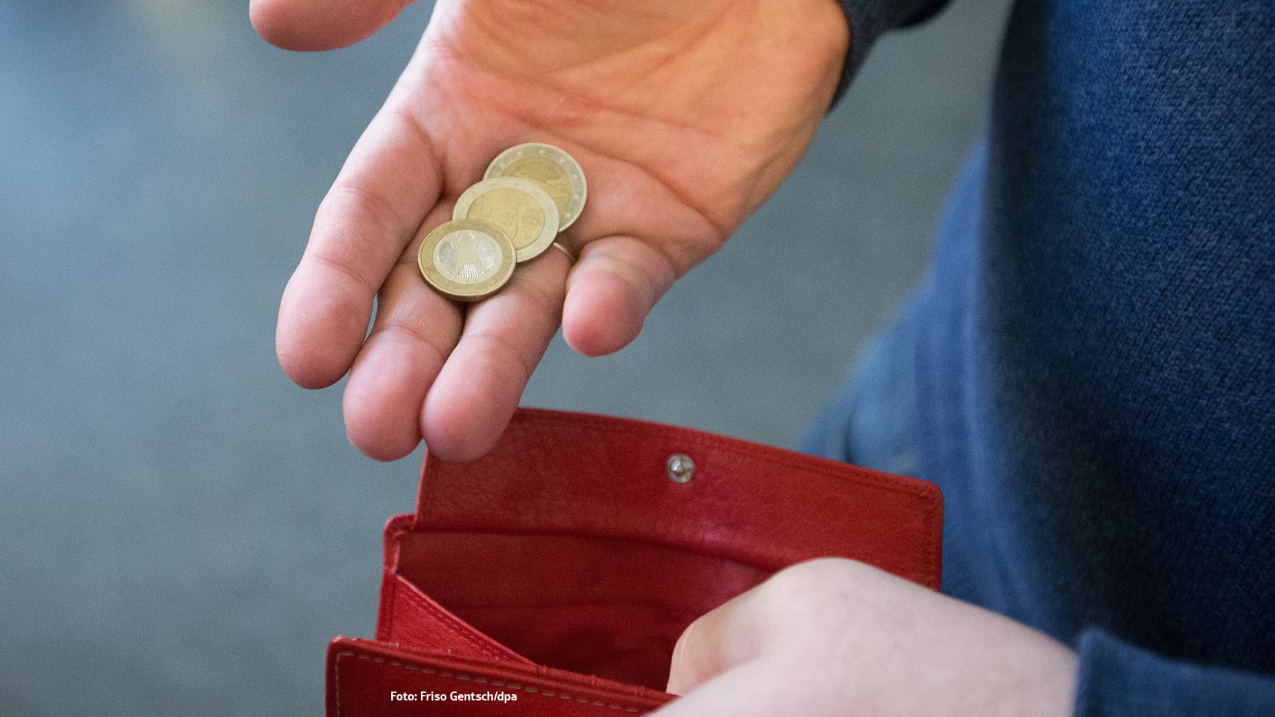 Eine Hand hält wenige Euromünzen über einer offenen roten Geldbörse, was Armut oder knappe Finanzen symbolisiert.