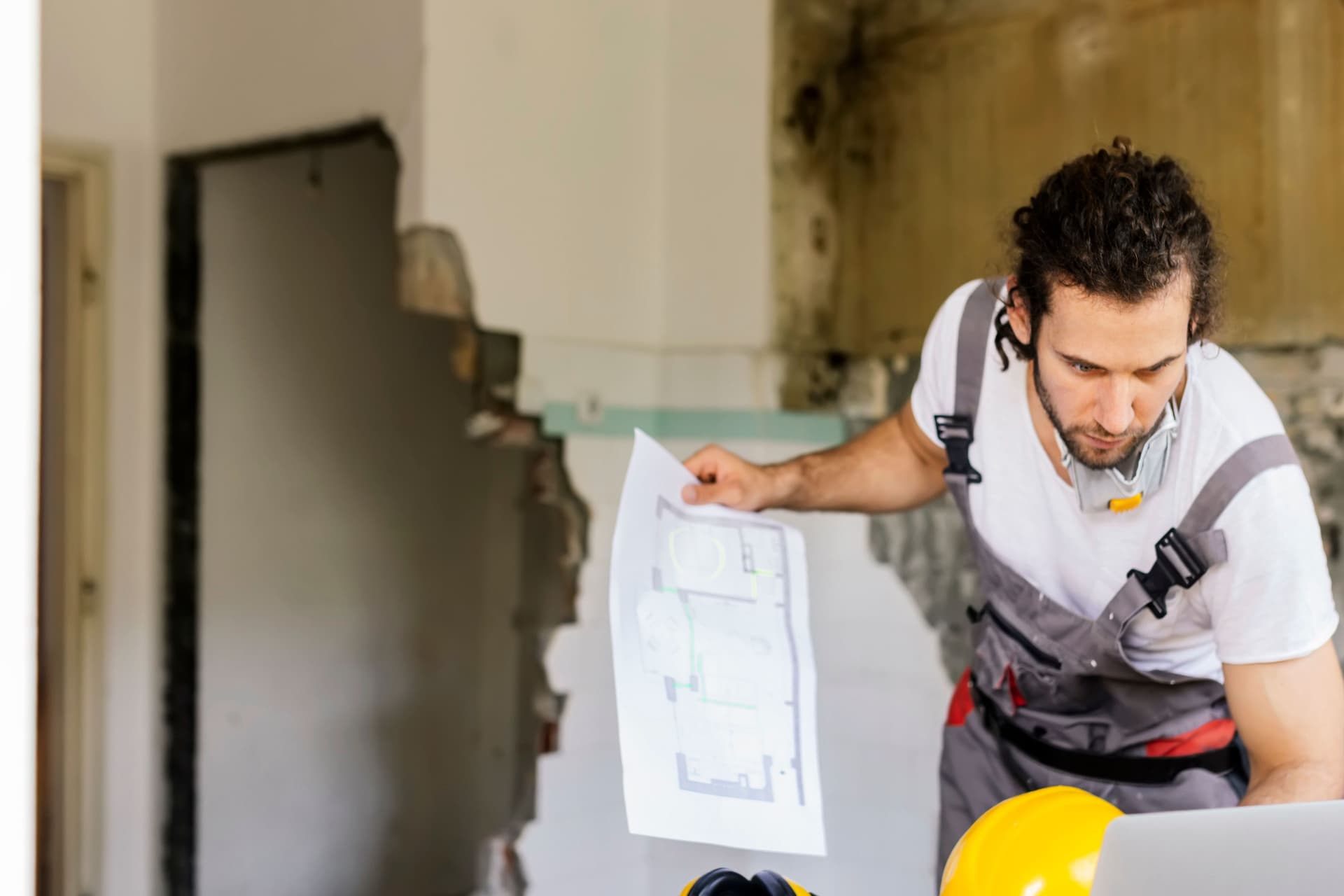 Arbeiter, der sich ein Architekturprojekt auf Papier und Laptop ansieht. Hinter ihm ist eine eingerissene Wand.