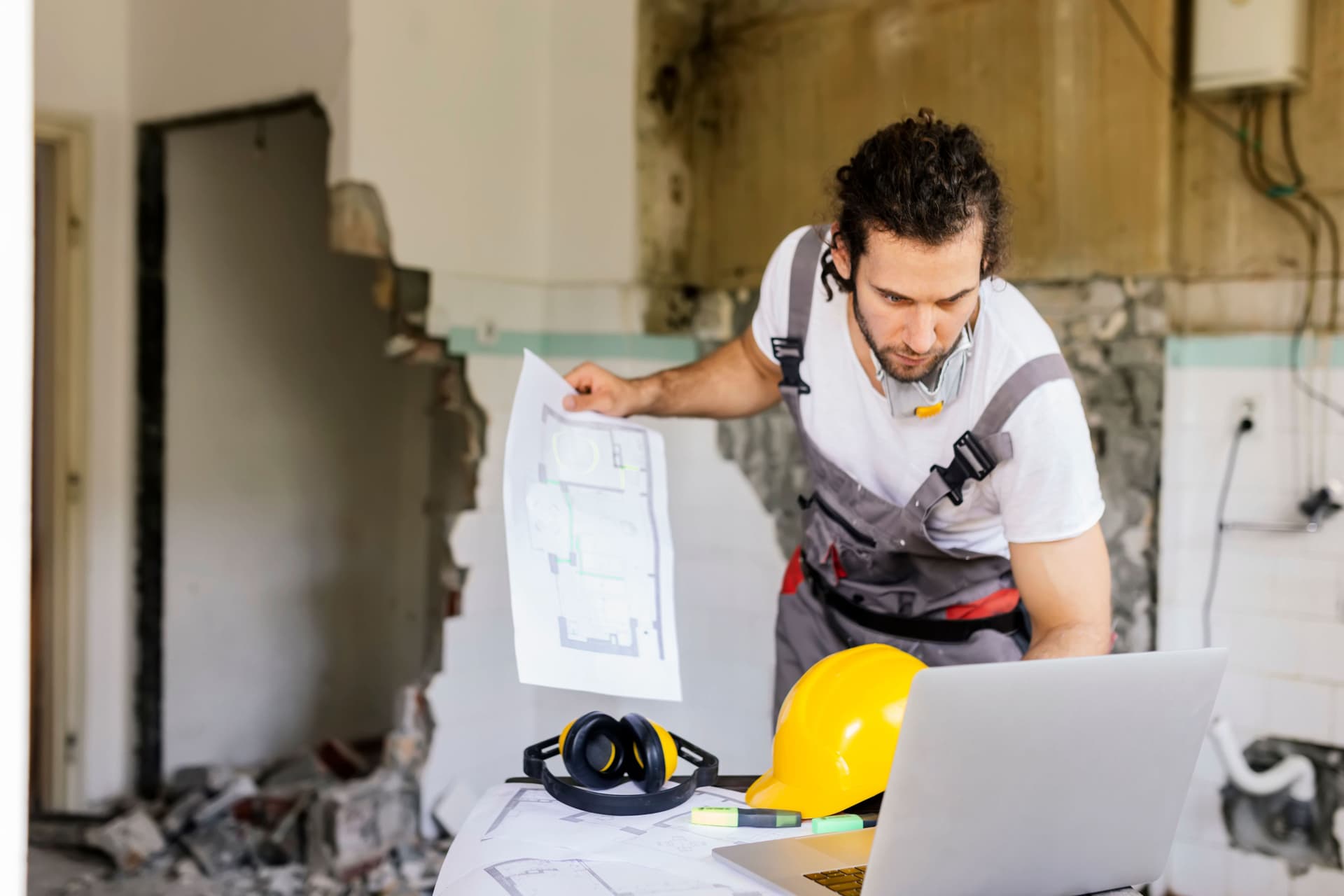 Arbeiter, der sich ein Architekturprojekt auf Papier und Laptop ansieht. Hinter ihm ist eine eingerissene Wand.
