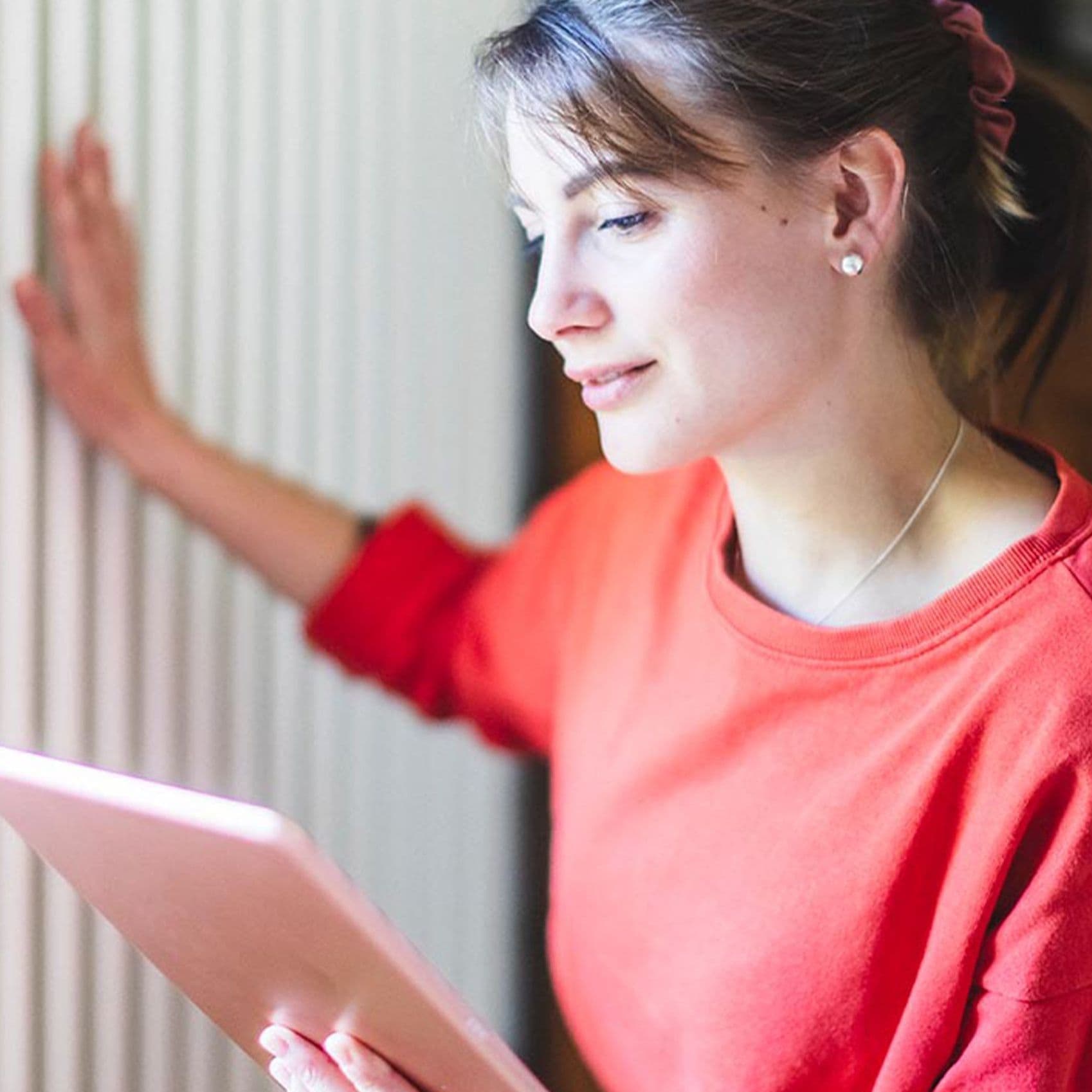 Eine junge Frau mit einem Tablet in der Hand fühlt die Temperatur eines Heizkörpers zuhause. 