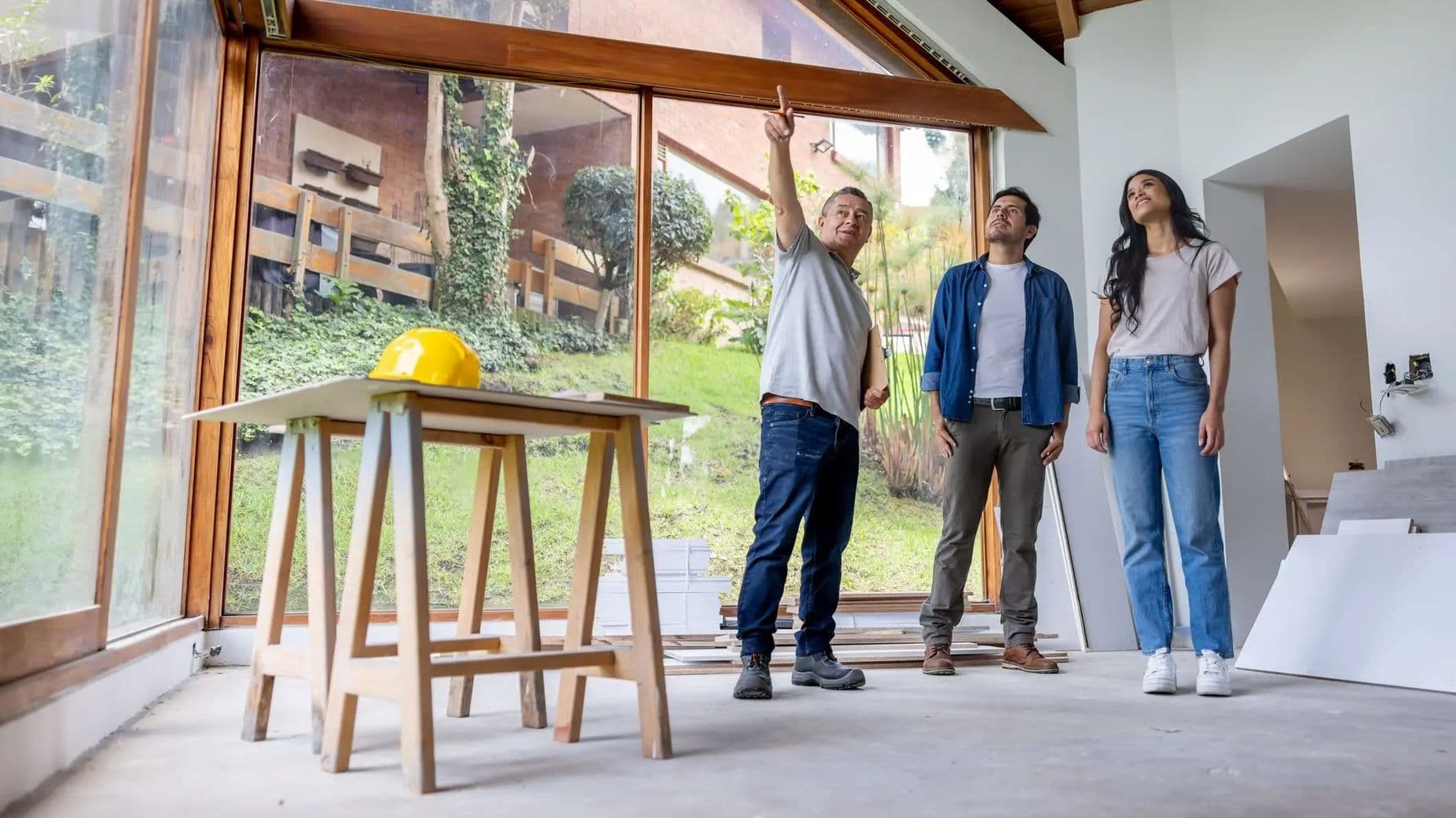 Architekt zeigt einem jungen Paar Details im Innenraum eines Neubaus mit großen Panoramafenstern und sichtbarem Holz-Dachstuhl