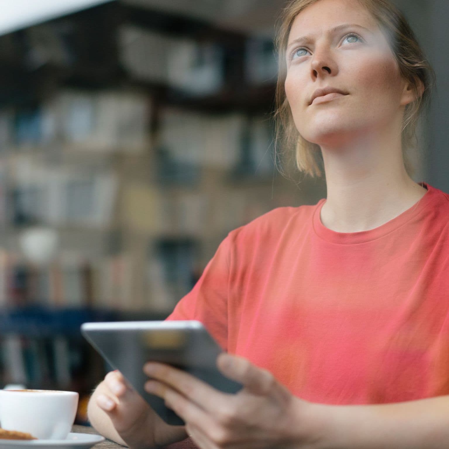 Eine junge Frau mit einem Tablet in der Hand sitzt in einem Café und schaut nach oben auf die Wohnhäuser gegenüber, die sich in der Fensterscheibe spiegeln. 