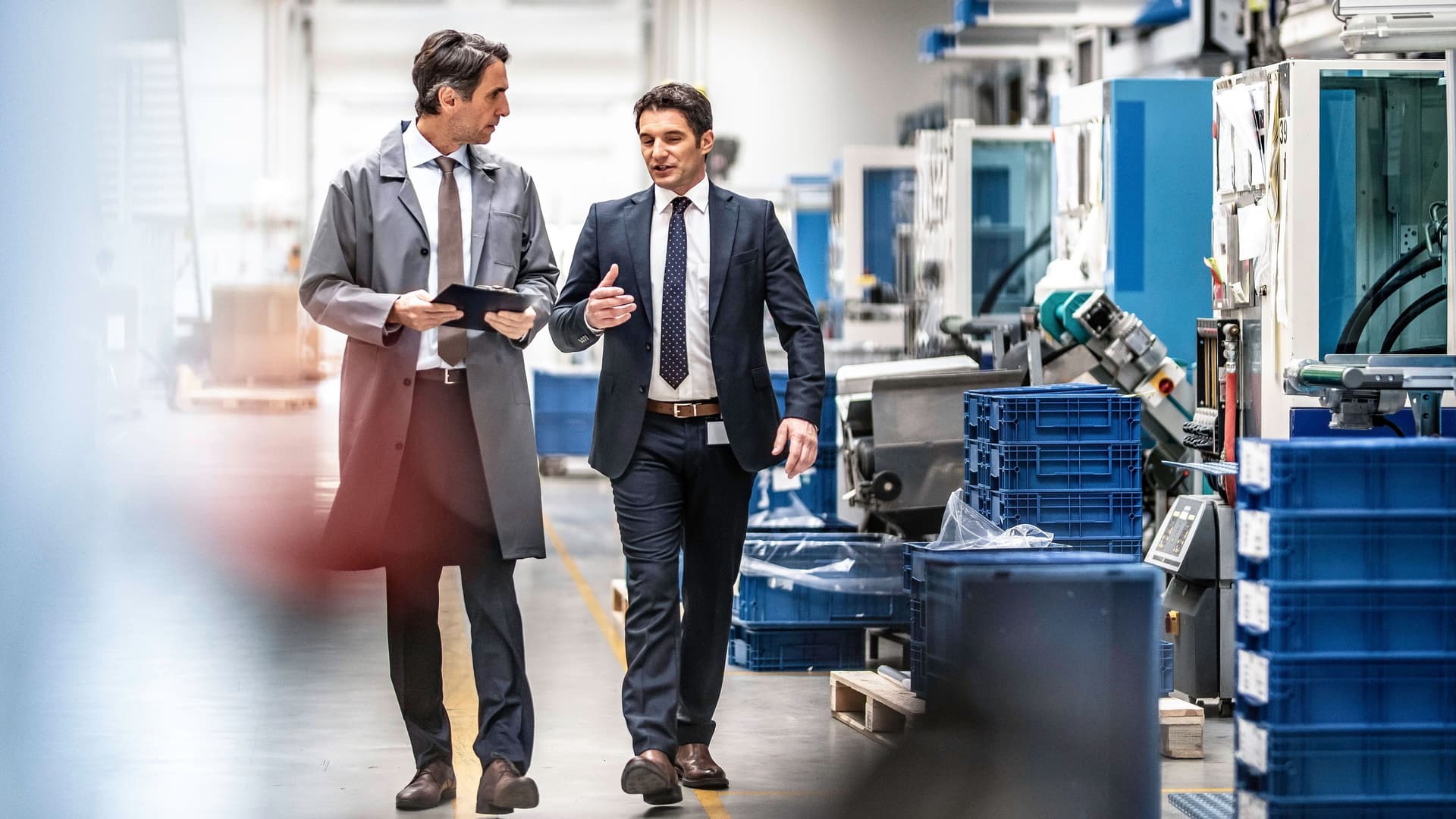 Ingenieur und Werkleiter laufen in Anzug durch eine Fabrik und reden miteinander.