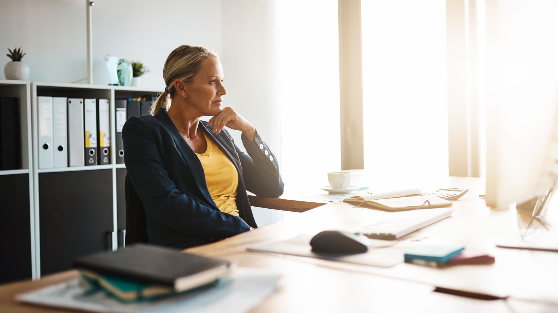 Eine Frau in einem Blazer sitzt an einem großen Bürotisch und stützt ihren Kopf nachdenklich auf ihre Hand. Die Sonne scheint durch ein großes Fenster in das Bürozimmer.