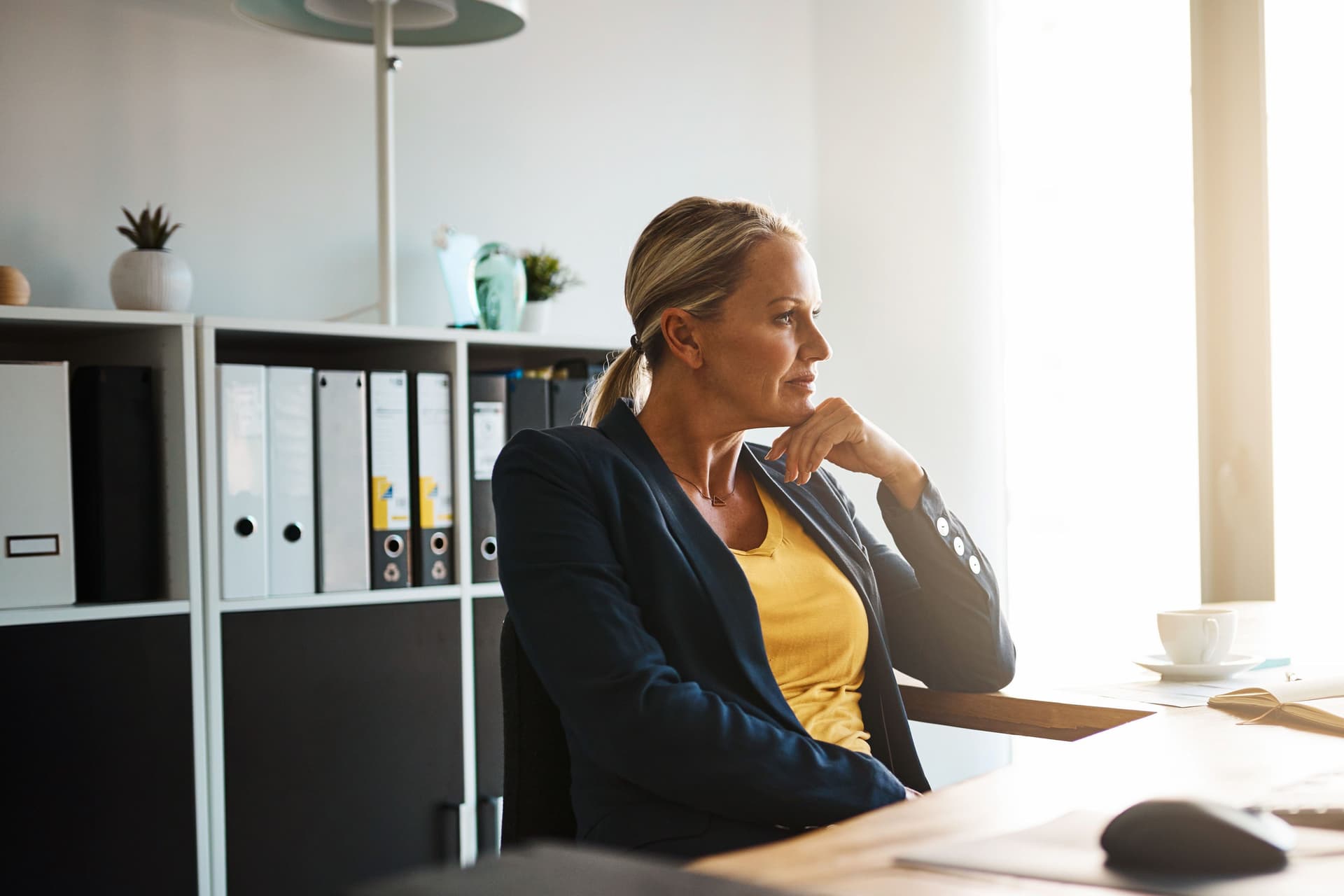 Eine Frau in einem Blazer sitzt an einem großen Bürotisch und stützt ihren Kopf nachdenklich auf ihre Hand. Die Sonne scheint durch ein großes Fenster in das Bürozimmer.