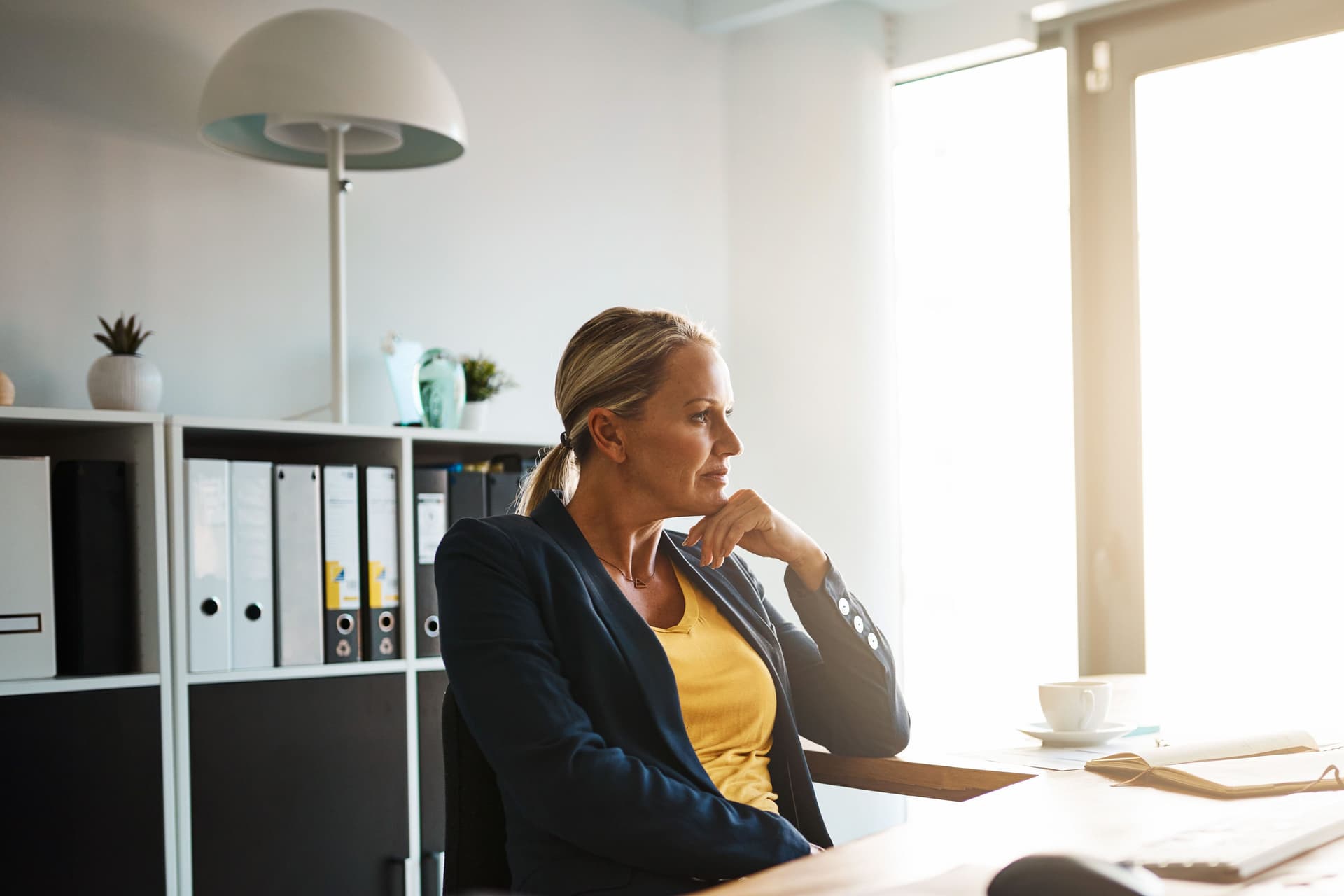 Eine Frau in einem Blazer sitzt an einem großen Bürotisch und stützt ihren Kopf nachdenklich auf ihre Hand. Die Sonne scheint durch ein großes Fenster in das Bürozimmer.