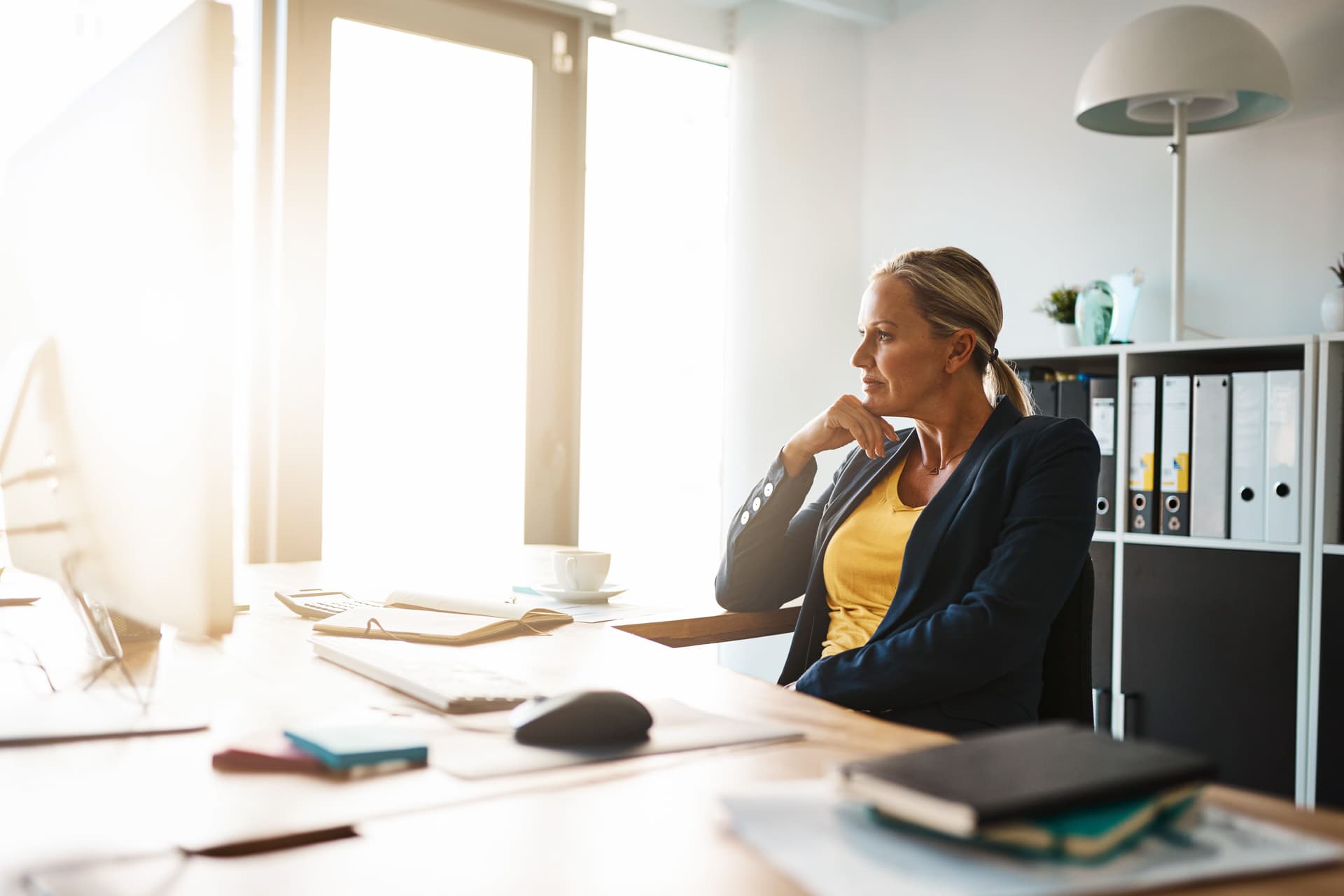 Eine Frau in einem Blazer sitzt an einem großen Bürotisch und stützt ihren Kopf nachdenklich auf ihre Hand. Die Sonne scheint durch ein großes Fenster in das Bürozimmer.