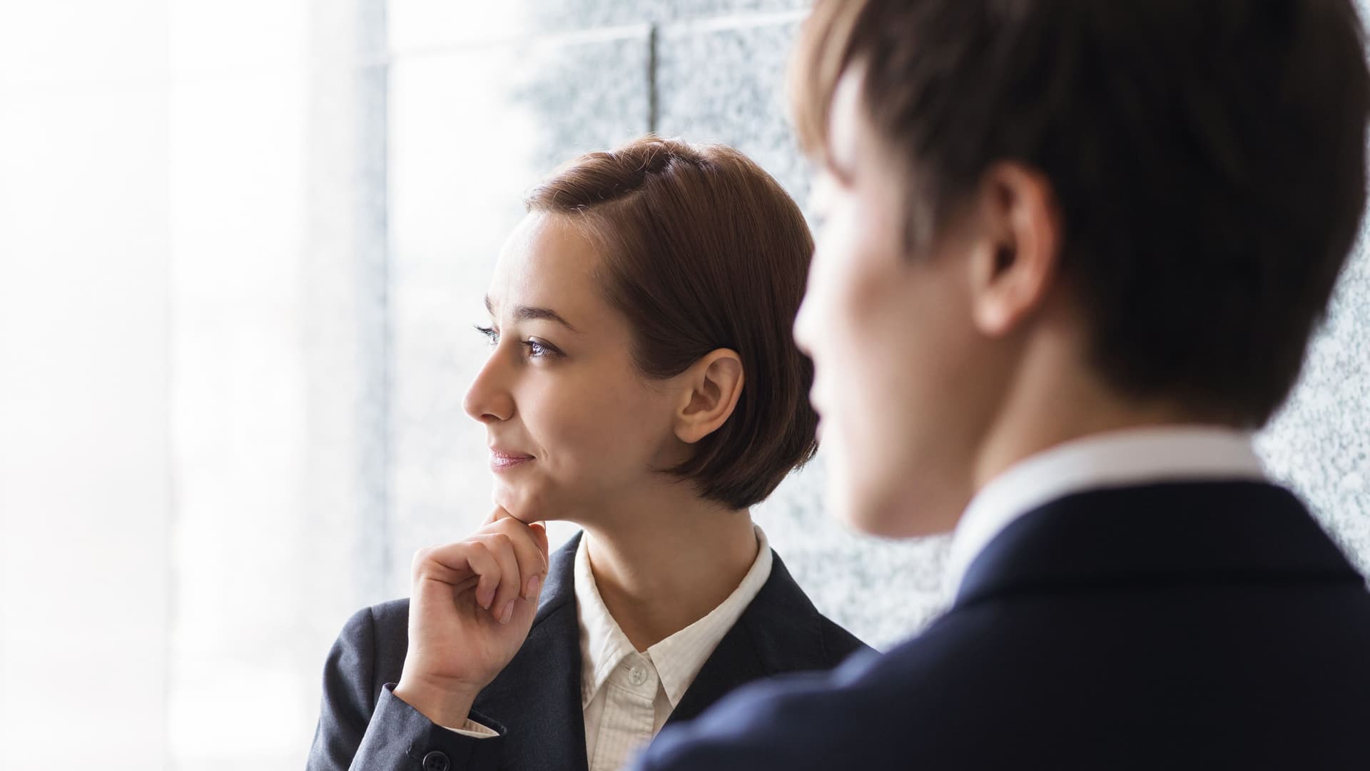 Junge Frau in Business-Kleidung hat ihre Hand am Kinn und guckt zur Seite. Vor ihr steht ein Mann in Anzug mit dem Rücken zur Kamera.