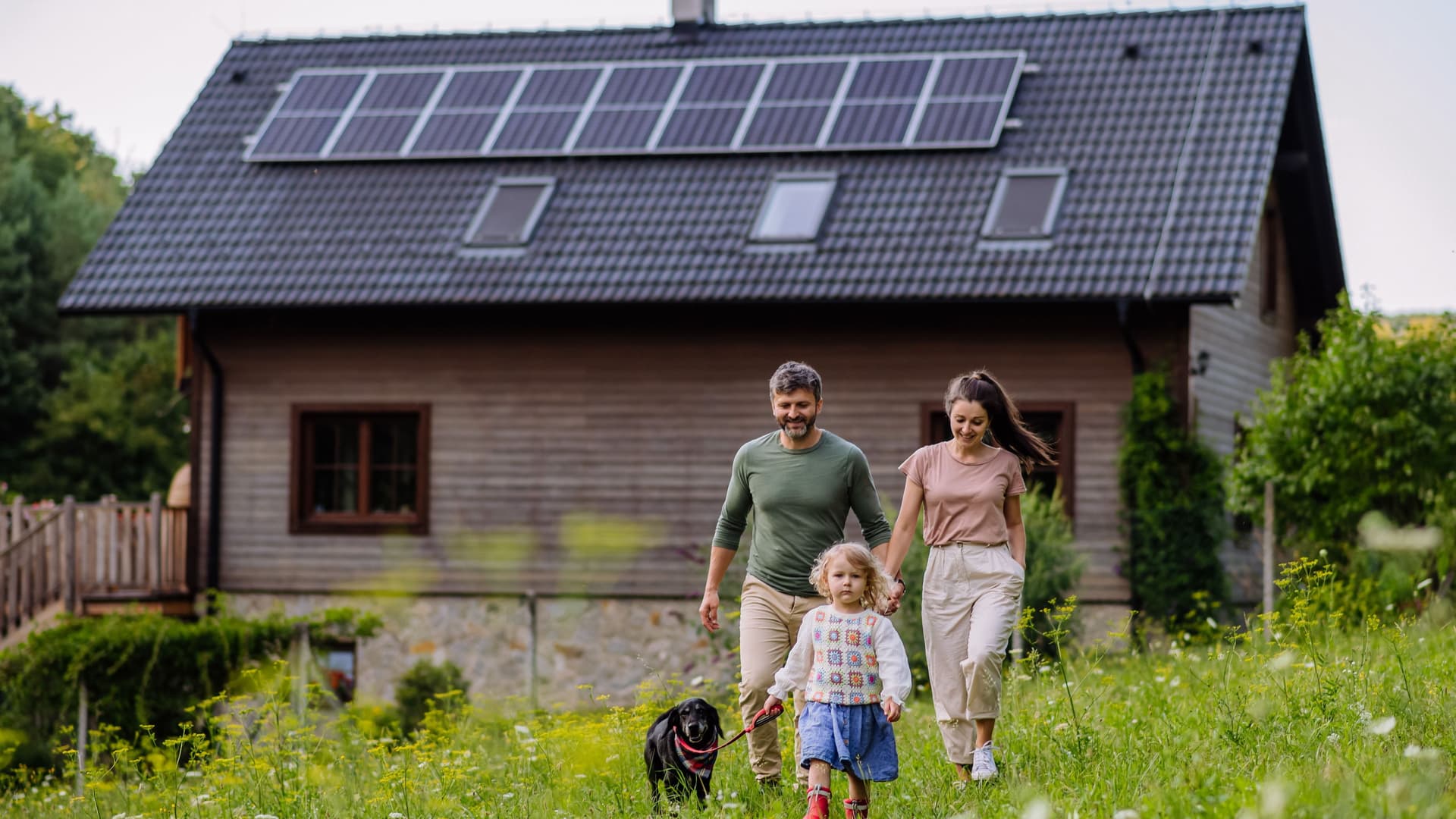 Junge Familie mit Tochter und Hund laufen über eine Wiese vor einem kleinen Haus mit Solarpanelen auf dem Dach.