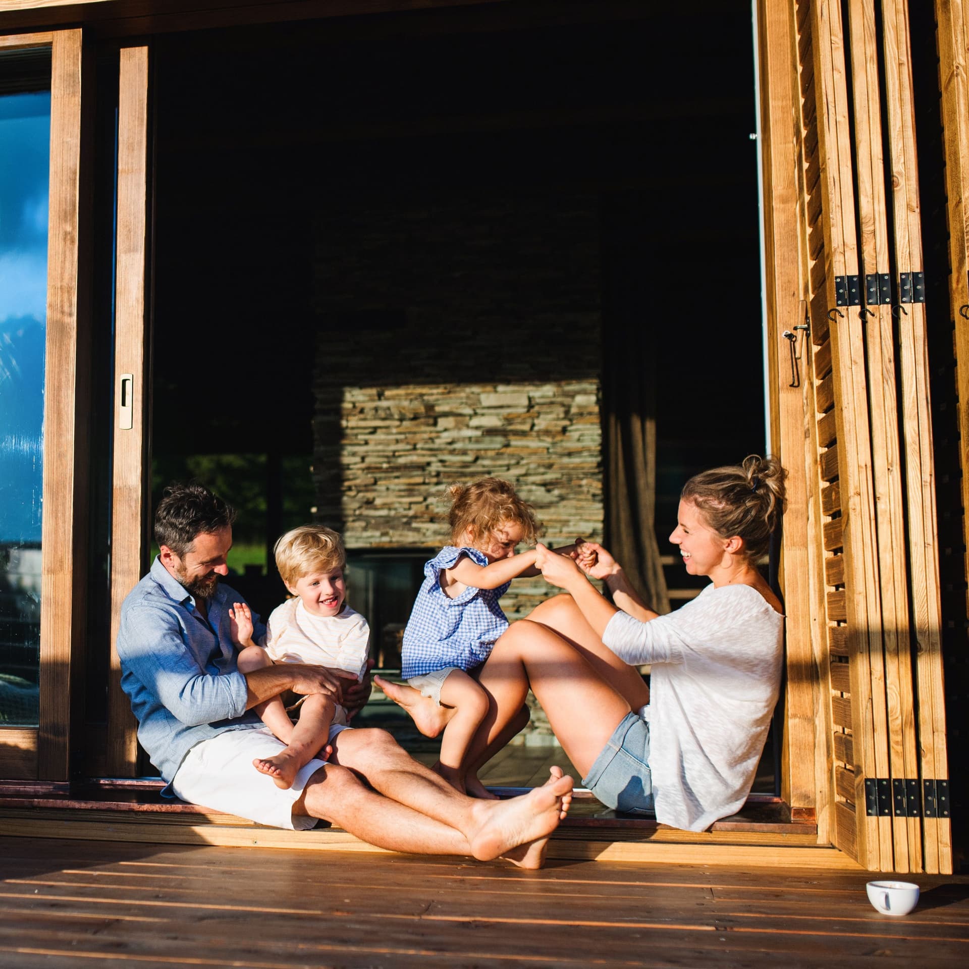 Familie mit kleinen Kindern, die auf dem Boden der Terrasse eines Holzhauses sitzen. Sie sind sommerlich gekleidet und die Sonne spiegelt sich in der Terassentür.