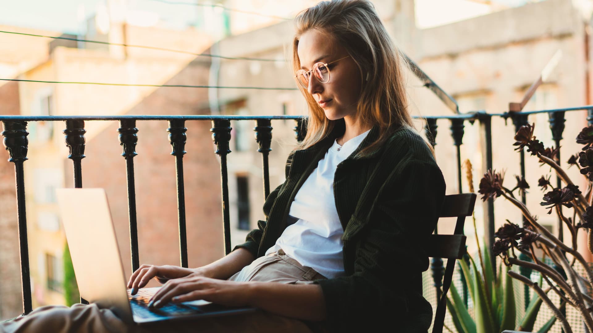 Junge Frau die von zu Hause aus am Laptop arbeitet und auf dem leicht sonnigen Balkon sitzt.