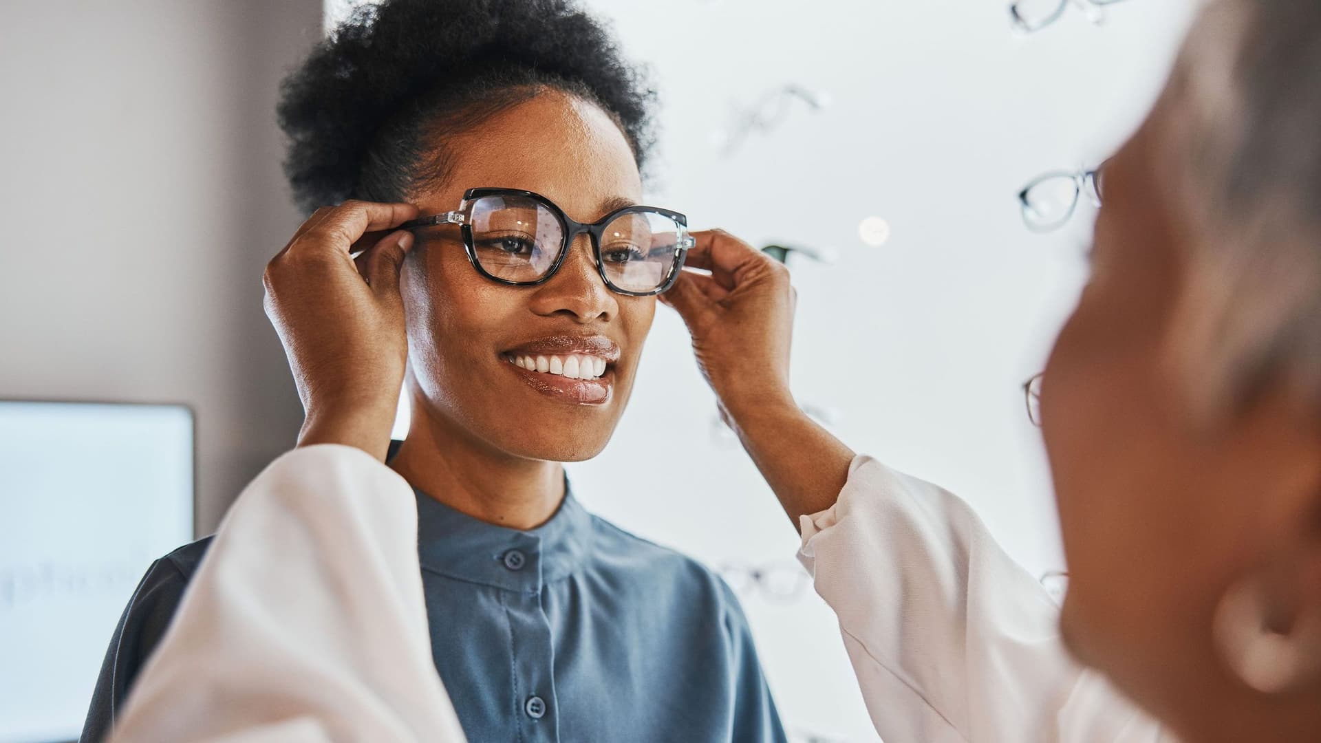 Eine junge Schwarze Frau probiert eine Brille und lächelt zufrieden. Eine Optikerin hilft ihr dabei, die Brille richtig anzupassen, während im Hintergrund weitere Brillen ausgestellt sind.