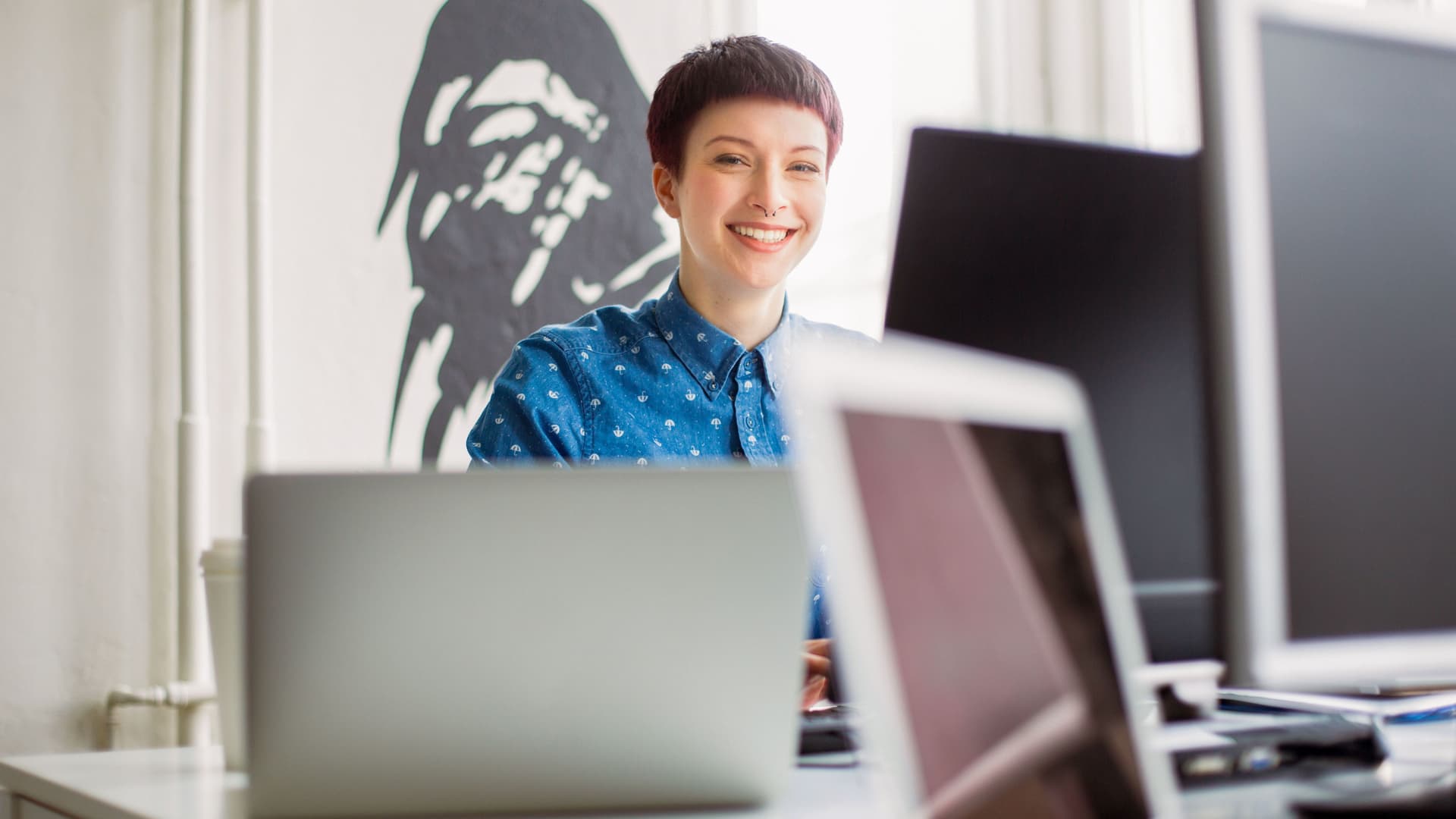 Junge Frau steht im Büro vor mehreren Monitoren und lächelt.