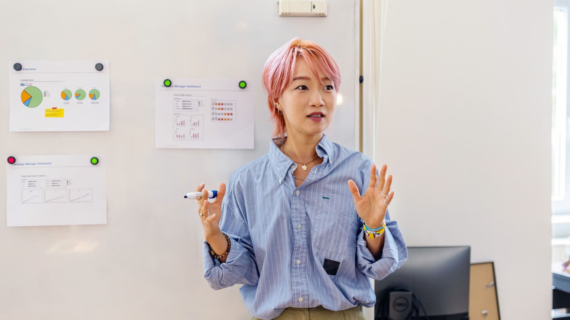 Junge Frau mit rosa gefärbten Haaren steht gestikulierend vor einem Whiteboard mit Charts und Graphen