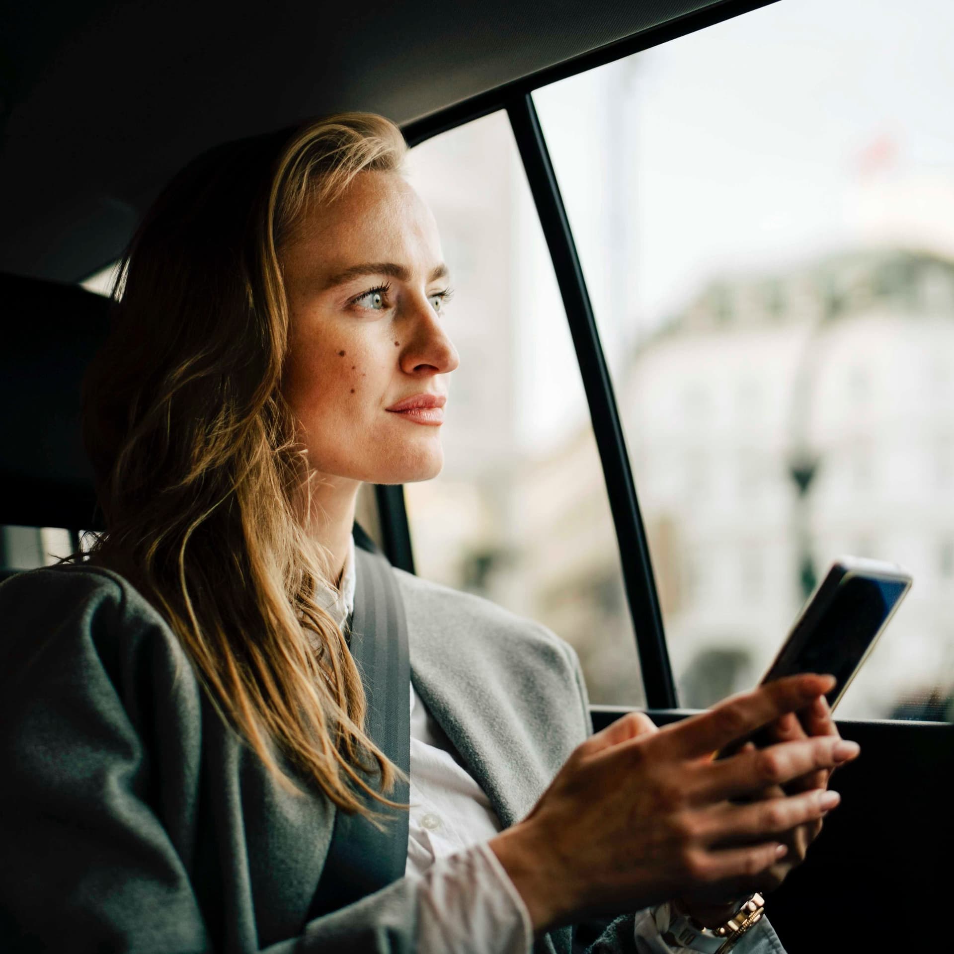Nachdenkliche junge Frau mit Smartphone in der Hand. Sie sitzt auf der Rückbank und schaut durch das Autofenster in die Landschaft.