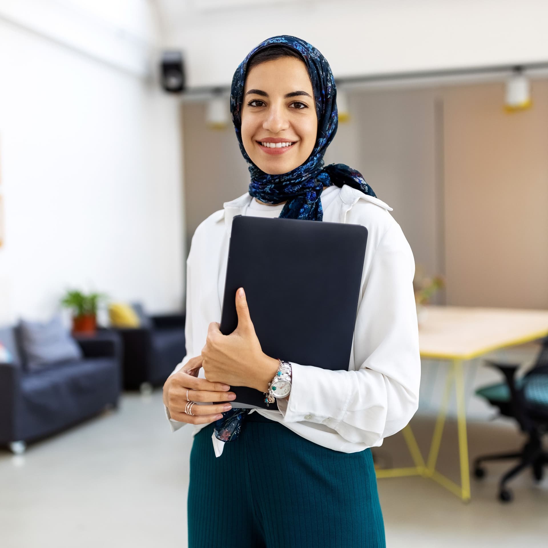 Eine junge Frau mit Hijab steht in einem großen Büro. Sie hält Unterlagen in einem Ordner in der Hand.
