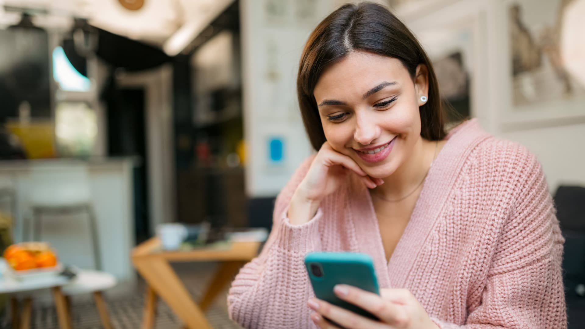 Junge Frau in rosa Strickjacke sitzt in einem Café und blickt lächelnd auf ihr Smartphone