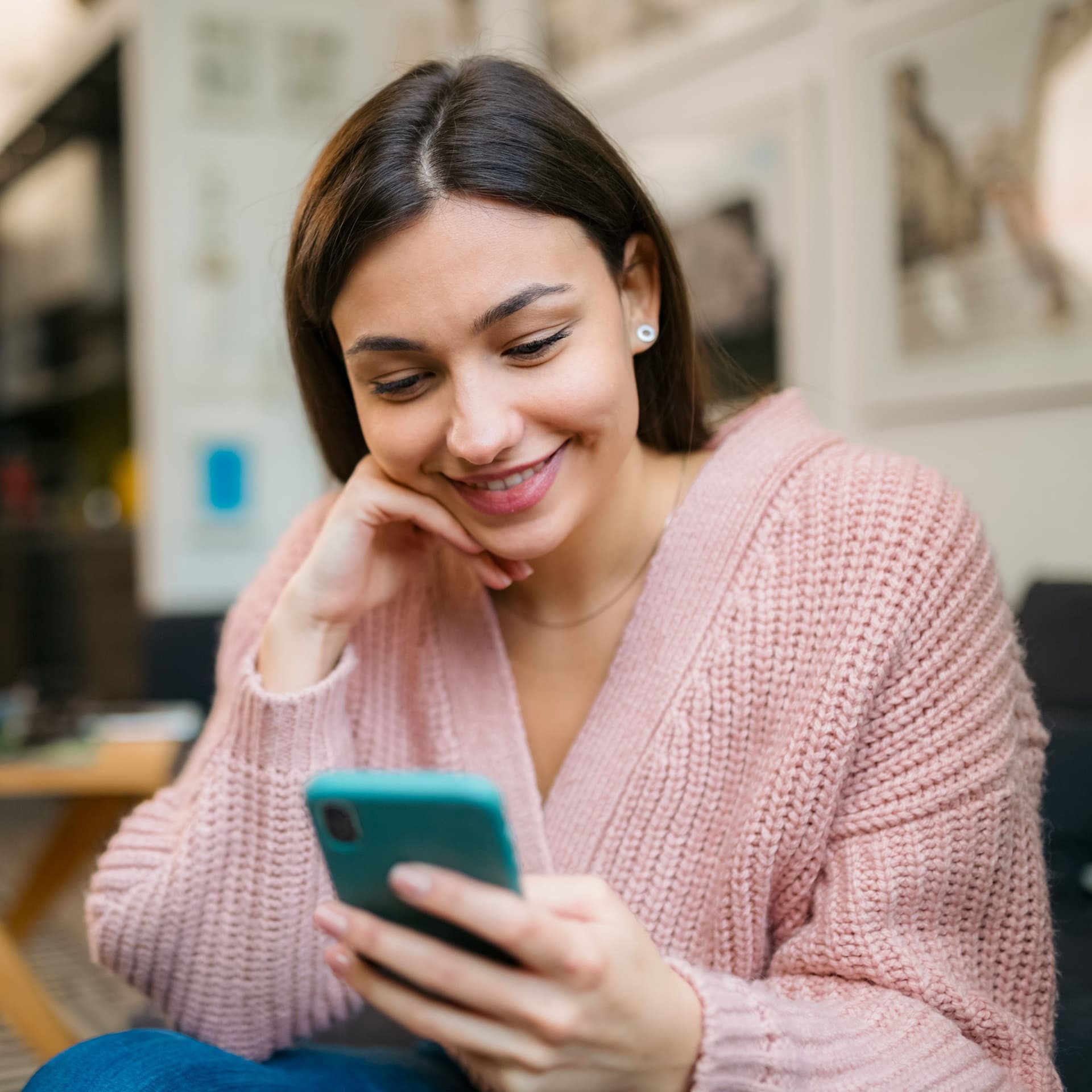 Junge Frau in rosa Strickjacke sitzt in einem Café und blickt lächelnd auf ihr Smartphone