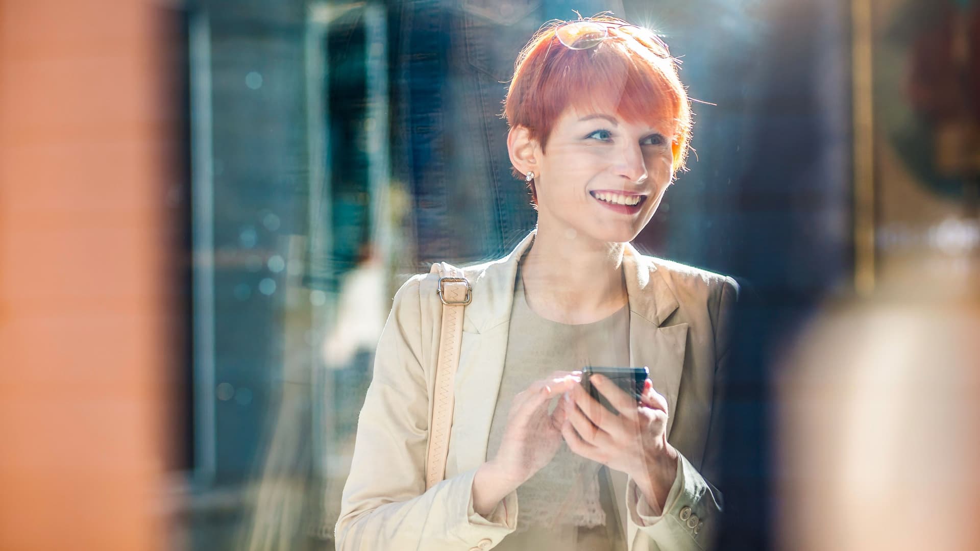 Blick durch ein Fenster auf eine lächelnde junge Frau mit roten Haaren, die ein Smartphone in der Hand hält.