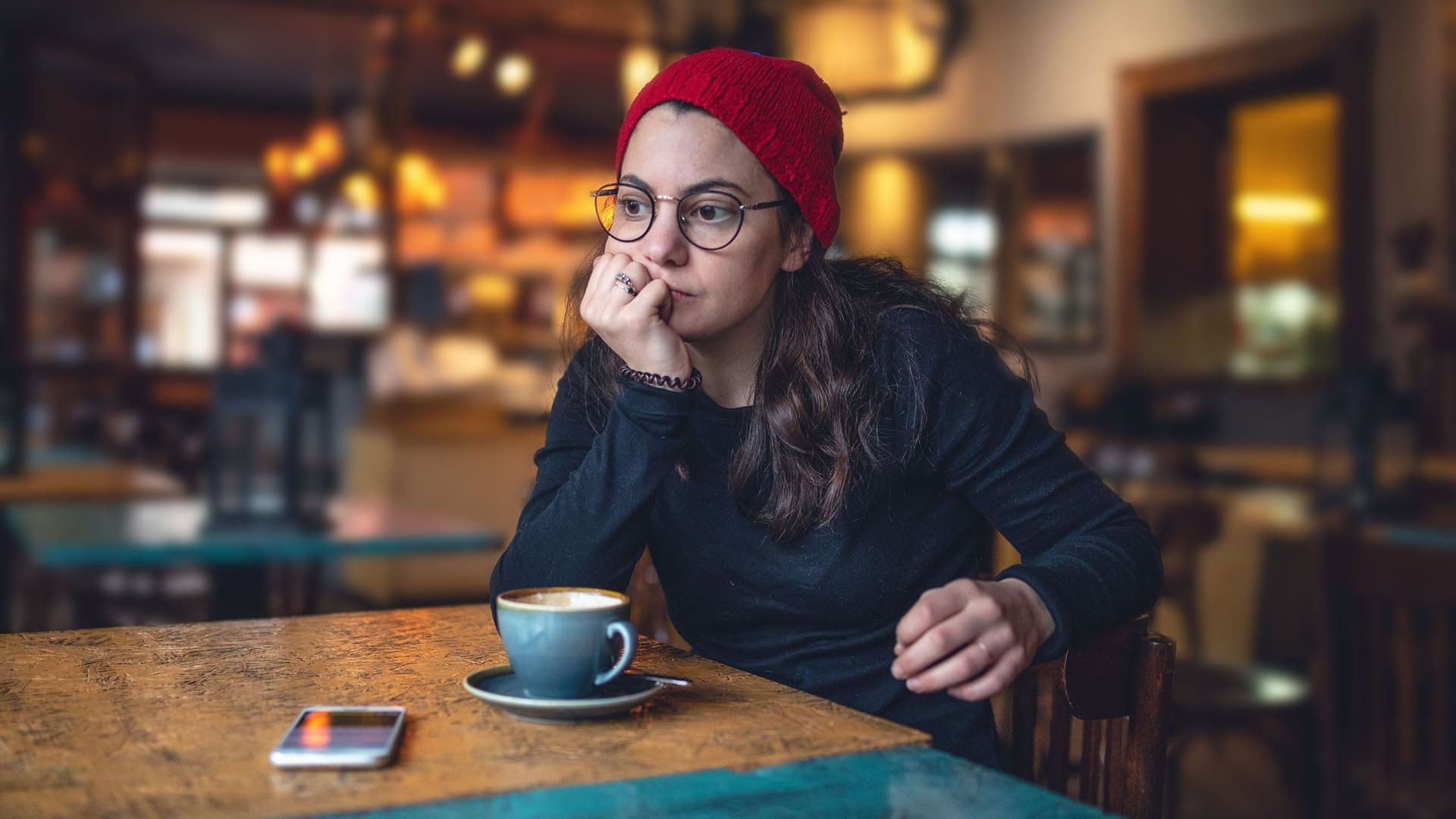 Eine junge Frau mit roter Mütze und runder Brille hat den Kopf auf die Hand gestützt und schaut ernst zum Cafe-Fenster. Vor ihr steht eine Kaffeetasse und ein Handy.