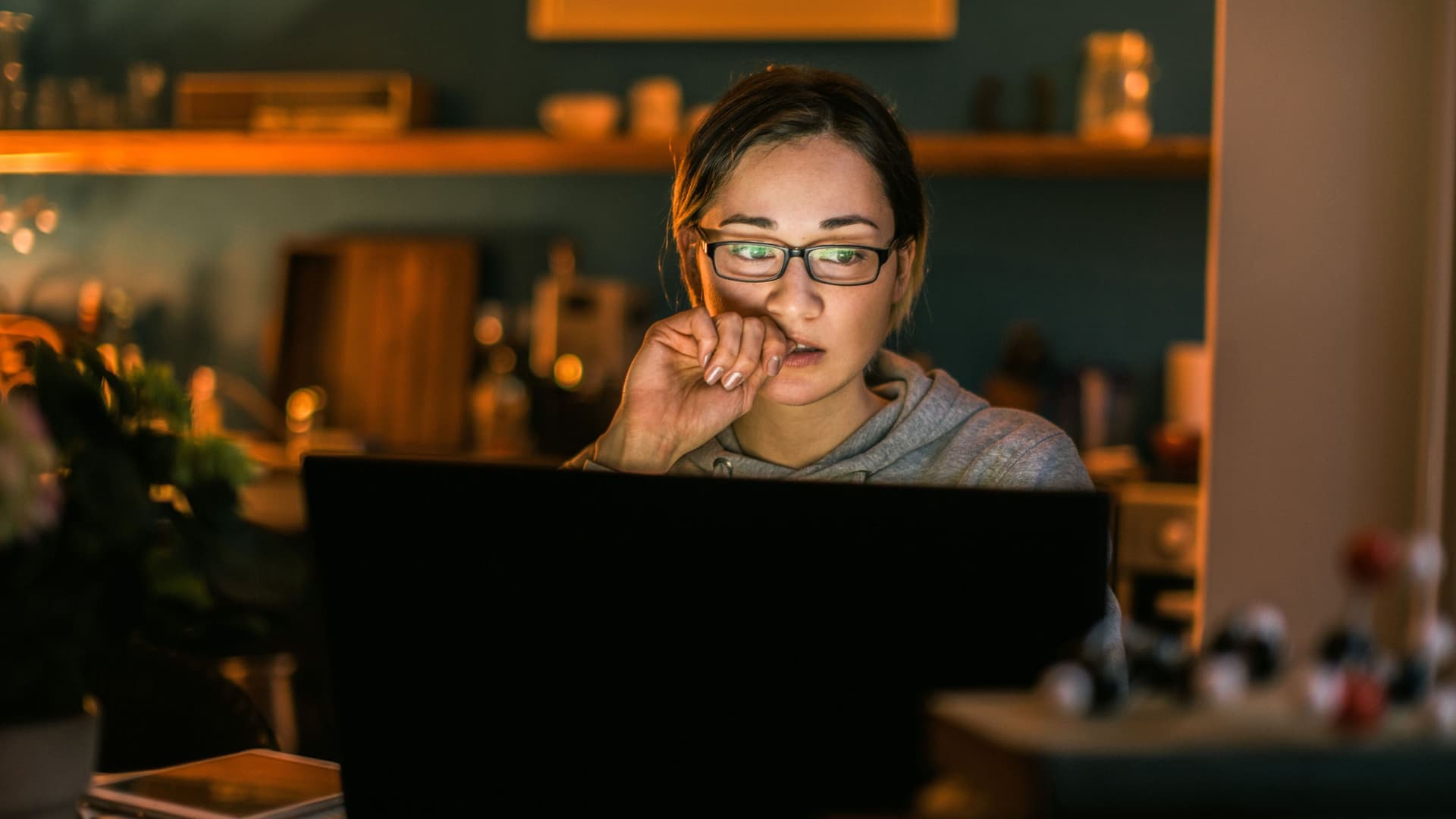 Junge Frau schaut  konzentriert auf ihren Laptop. Es ist Abend und ihre Wohnung ist dunkel.