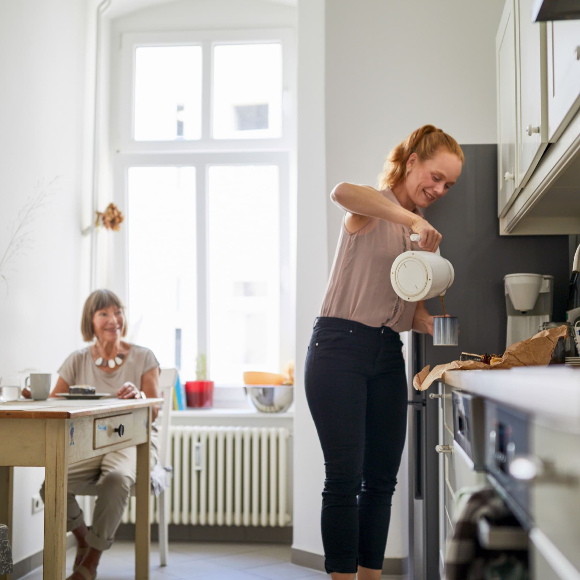 Junge Frau kocht Kaffee für eine Seniorin am Küchentisch.