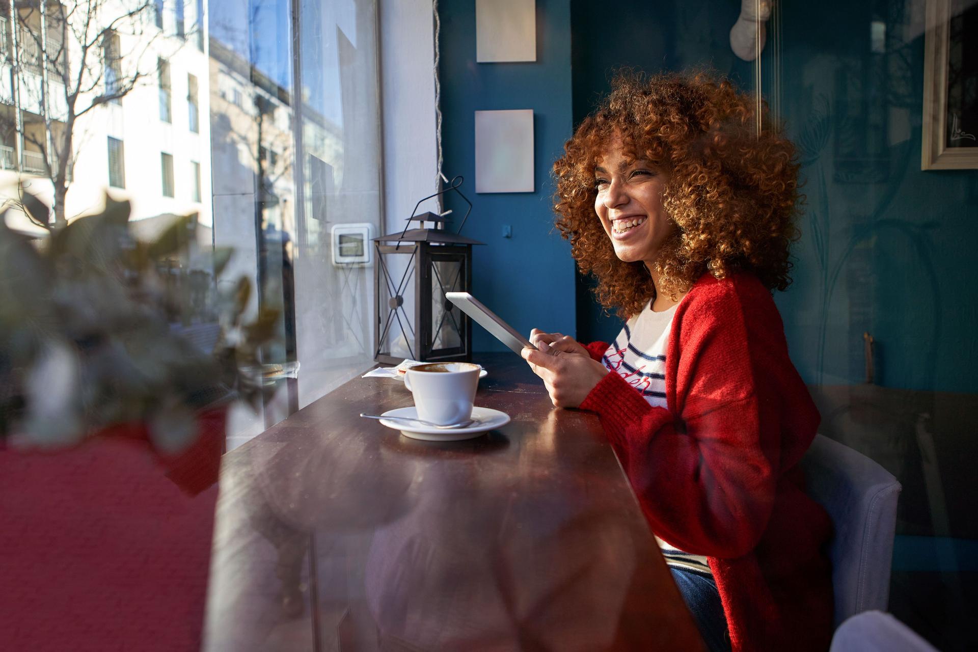 Eine junge Frau sitzt lachend im Cafe, blickt aus dem Fenster und hält ein Mobiltelefon in der Hand