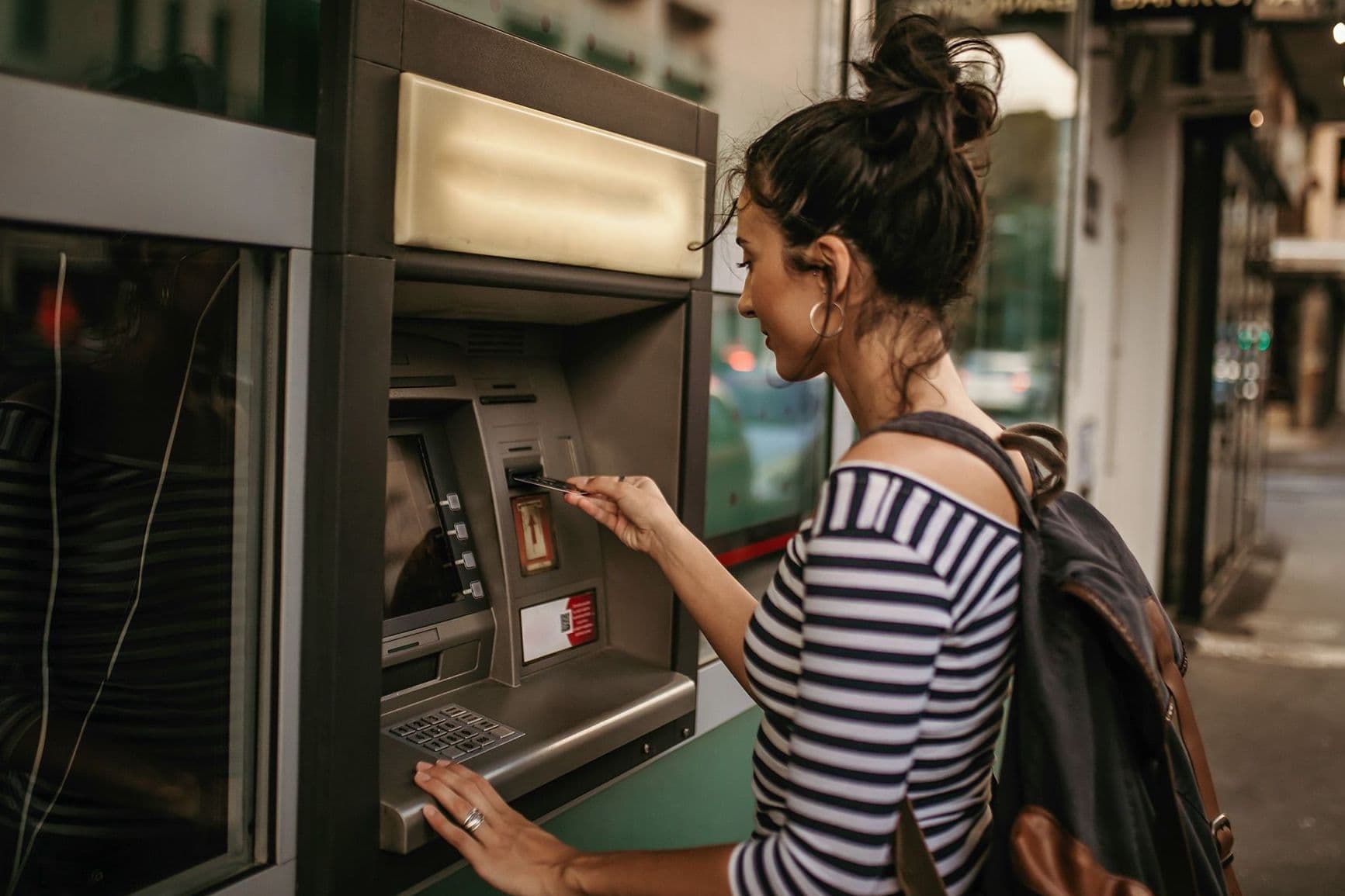 Eine junge Frau mit Dutt und Rucksack steht vor einem Straßen-Bankautomaten und führt ihre Bankkarte ein. 