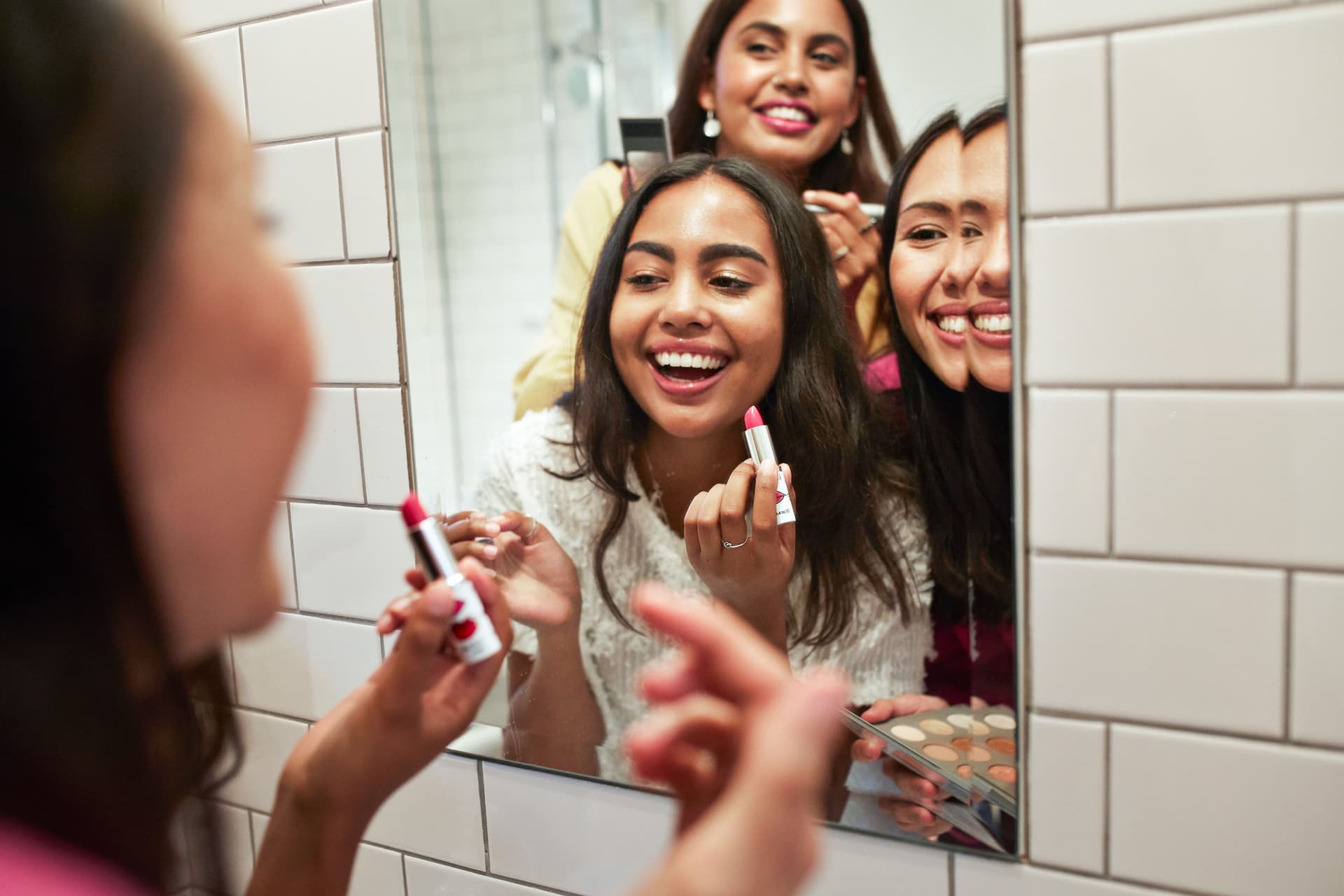 Drei junge Frauen lachen vor einem Spiegel an einer gefliesten Wand, eine hält einen Lippenstift in der Hand
