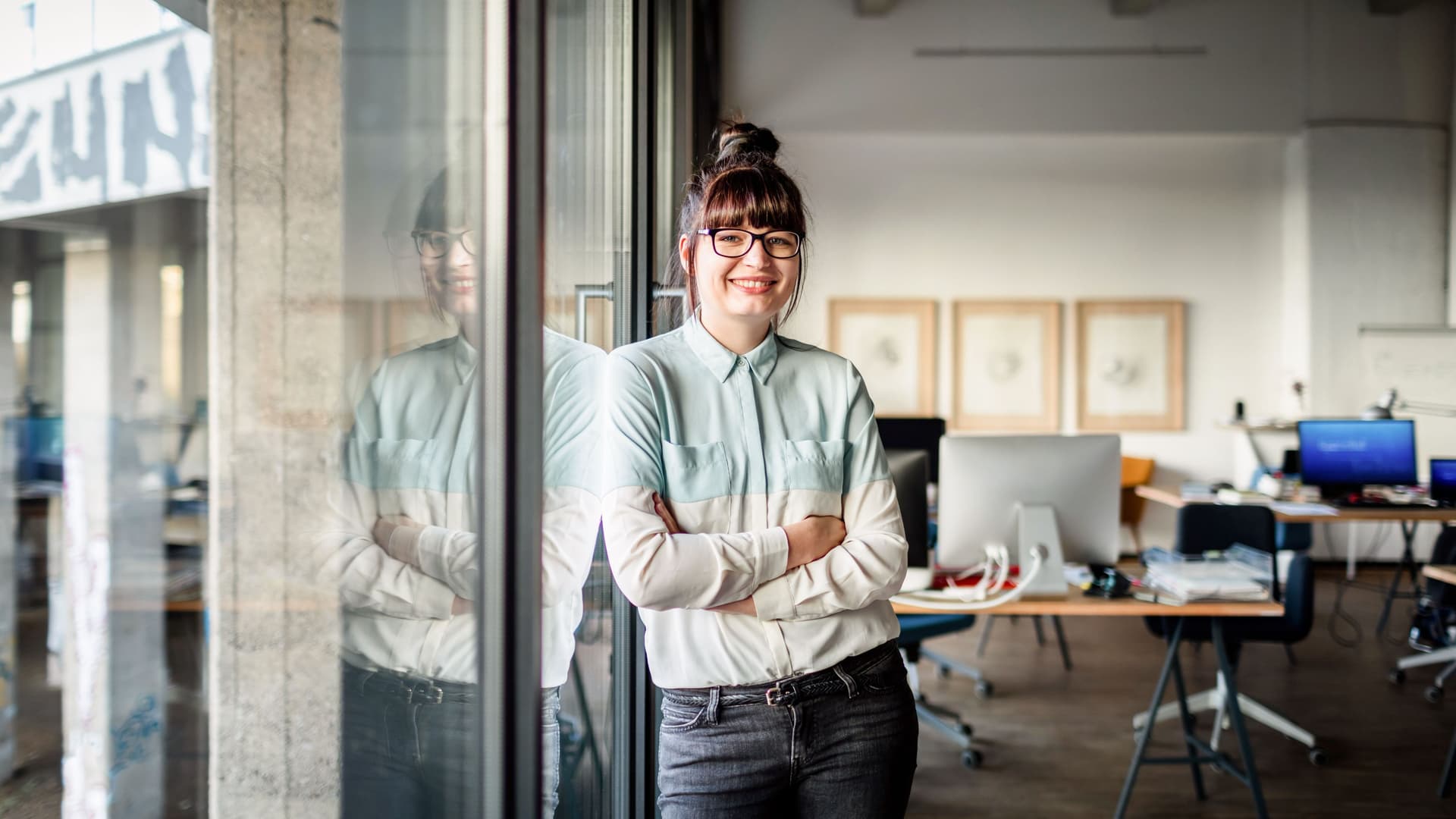 Junge Frau mit Brille und Dutt, die mit verschränkten Armen an einer Glasscheibe in einem modernen Büro lehnt und in die Kamera lächelt.