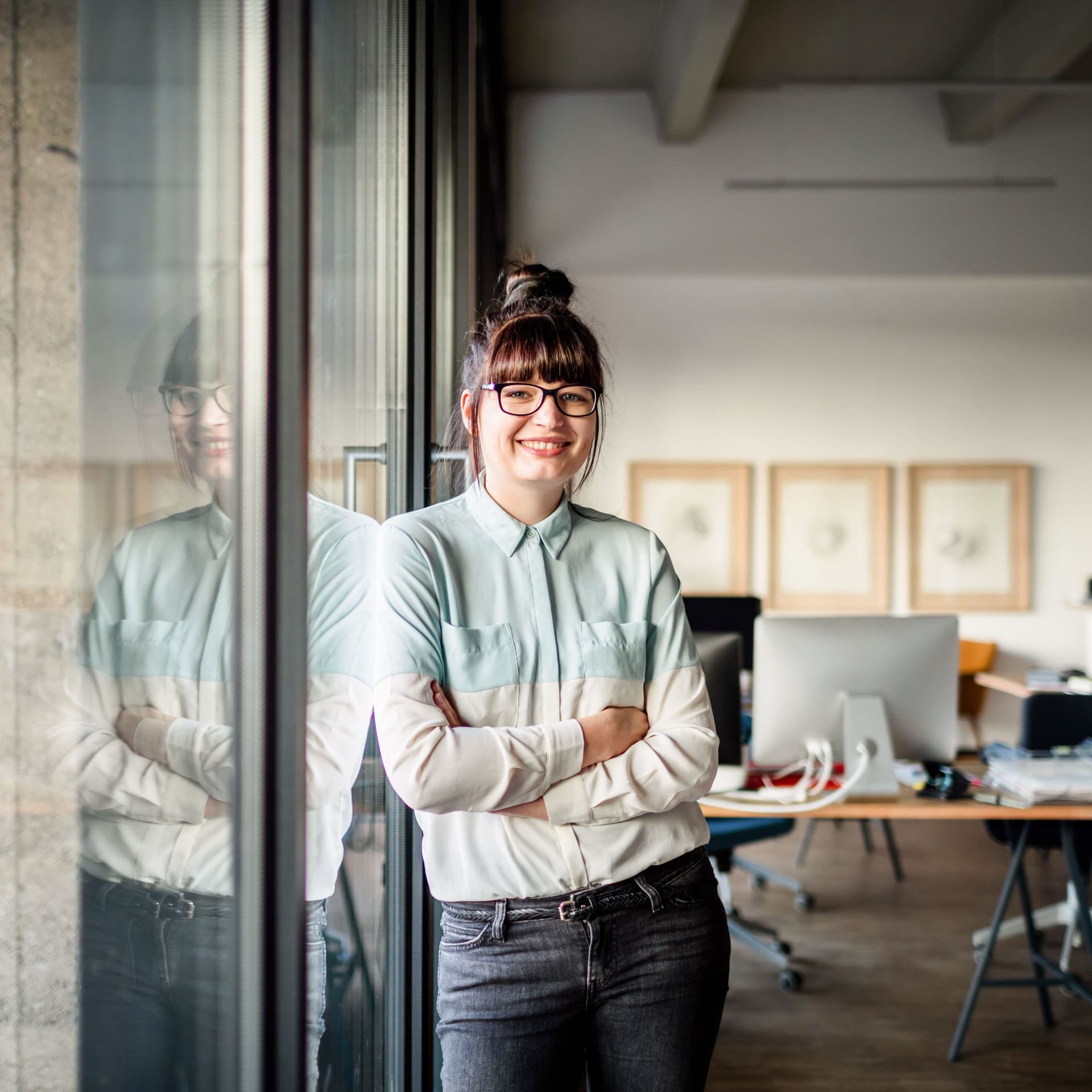 Junge Frau mit Brille und Dutt, die mit verschränkten Armen an einer Glasscheibe in einem modernen Büro lehnt und in die Kamera lächelt.