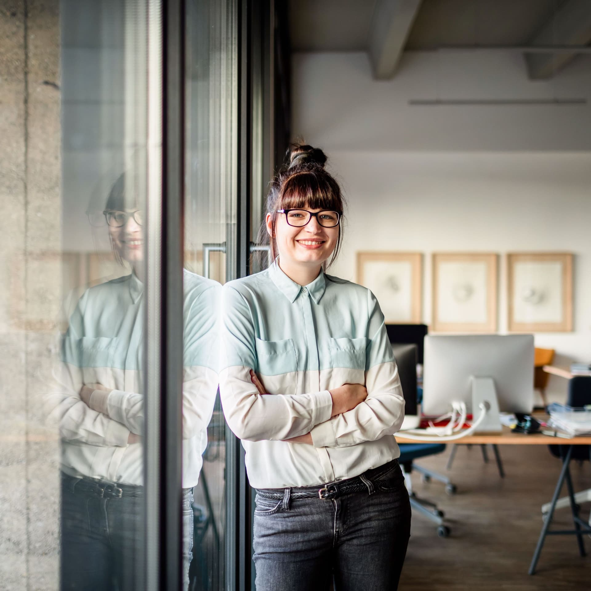 Junge Frau mit Brille und Dutt, die mit verschränkten Armen an einer Glasscheibe in einem modernen Büro lehnt und in die Kamera lächelt.