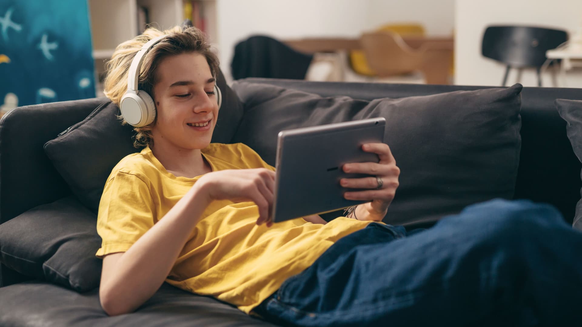 Junge liegt gemütlich auf dem Sofa. Er trägt Kopfhörer und hält lächelnd ein Tablet.