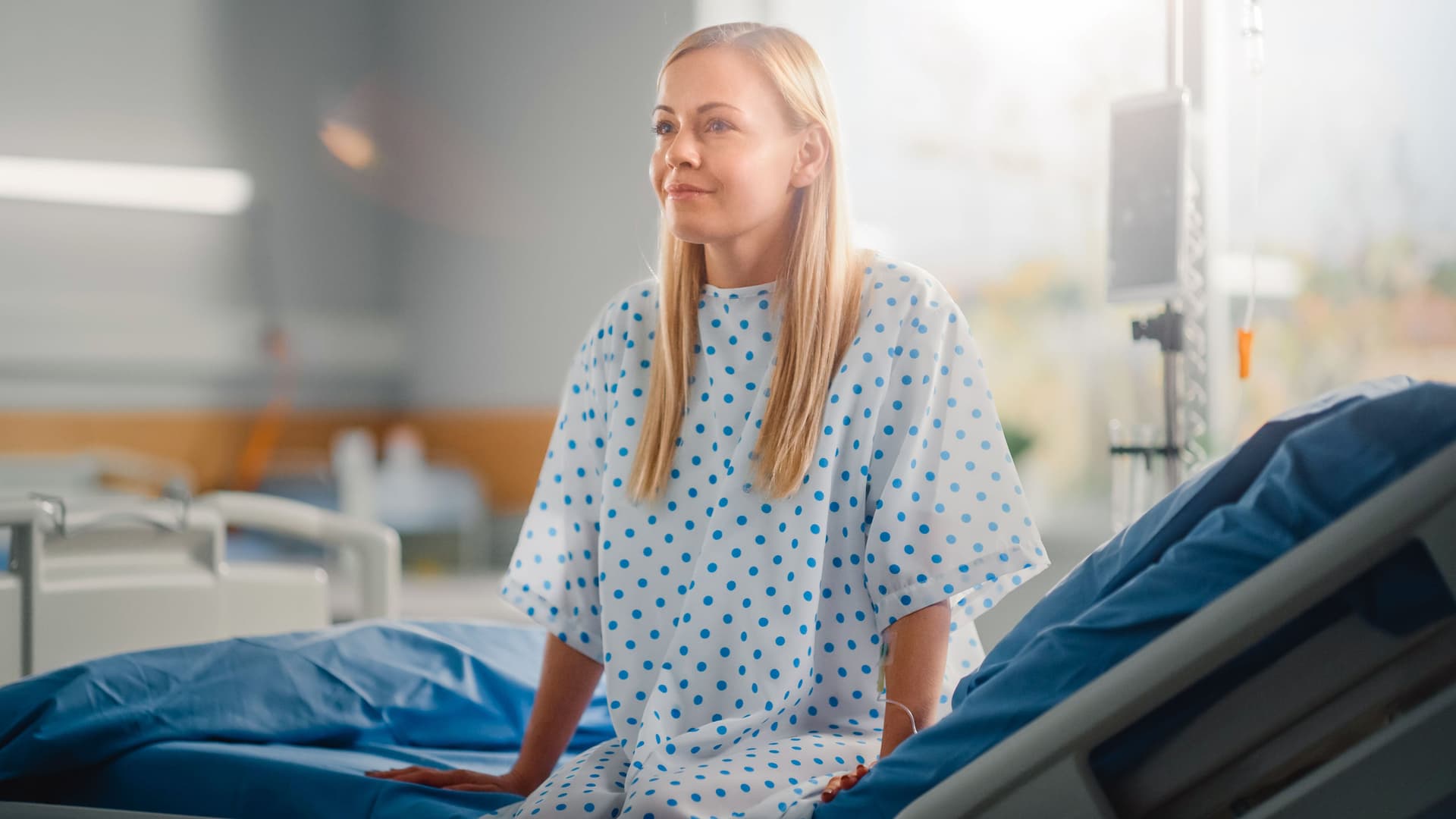 Eine Frau sitzt im Krankenhaus auf einem Bett in einem Krankenhaushemd. Sie lächelt dabei. Im Hintergrund sind weitere Betten und ein Glasfenster zu erkennen.
