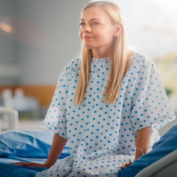 Eine Frau sitzt im Krankenhaus auf einem Bett in einem Krankenhaushemd. Sie lächelt dabei. Im Hintergrund sind weitere Betten und ein Glasfenster zu erkennen.