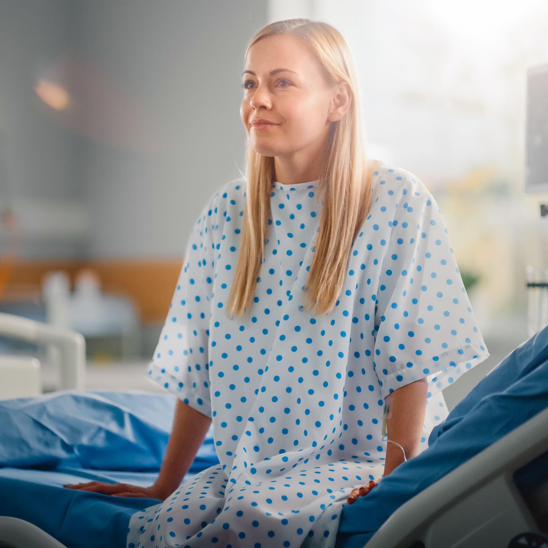 Eine Frau sitzt im Krankenhaus auf einem Bett in einem Krankenhaushemd. Sie lächelt dabei. Im Hintergrund sind weitere Betten und ein Glasfenster zu erkennen.