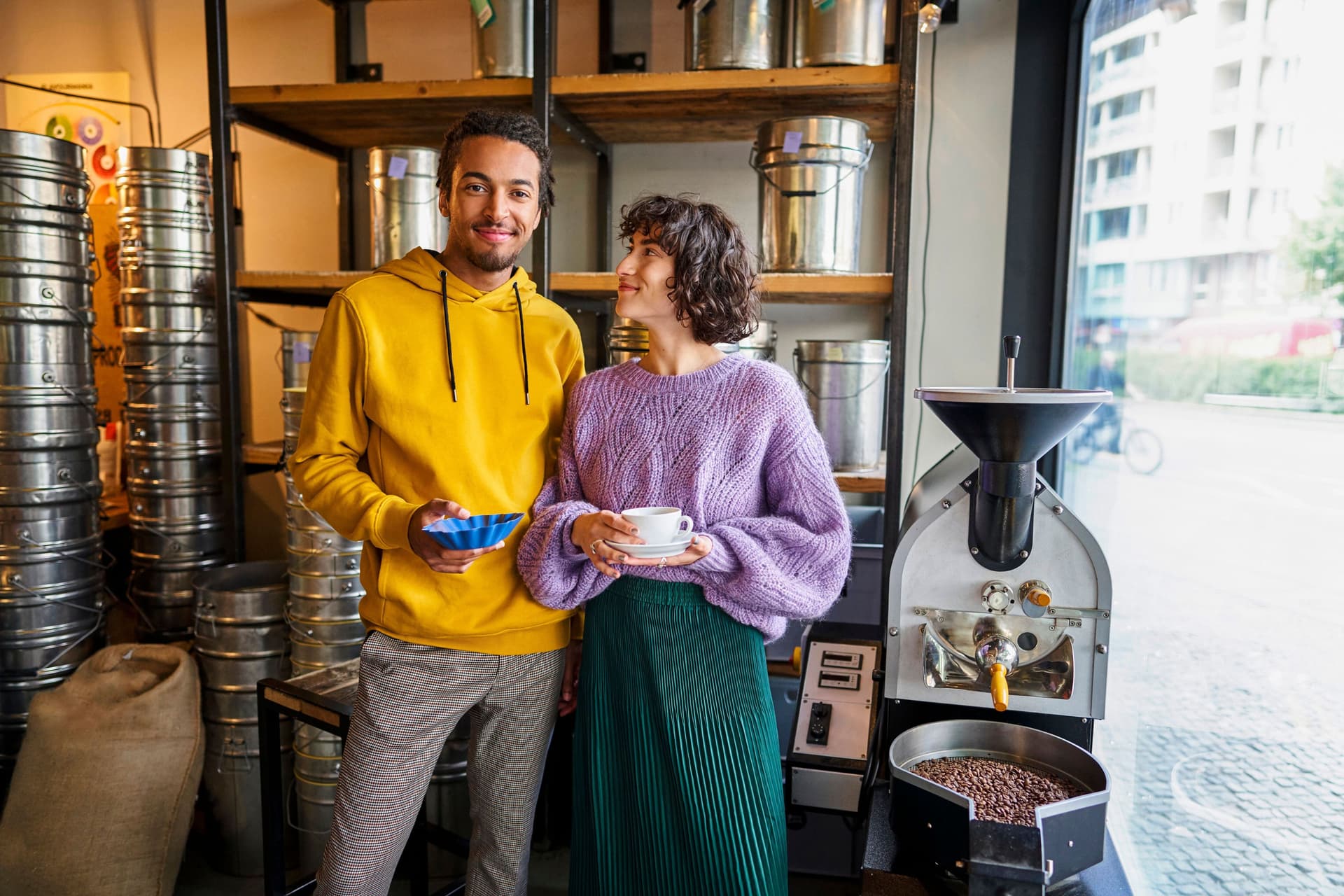 Eine junge Frau und ein junger Mann stehen in einer Kaffeerösterei in einem Ladengeschäft. Sie hat eine Tasse in der Hand und er eine Schale mit Kaffeebohnen.