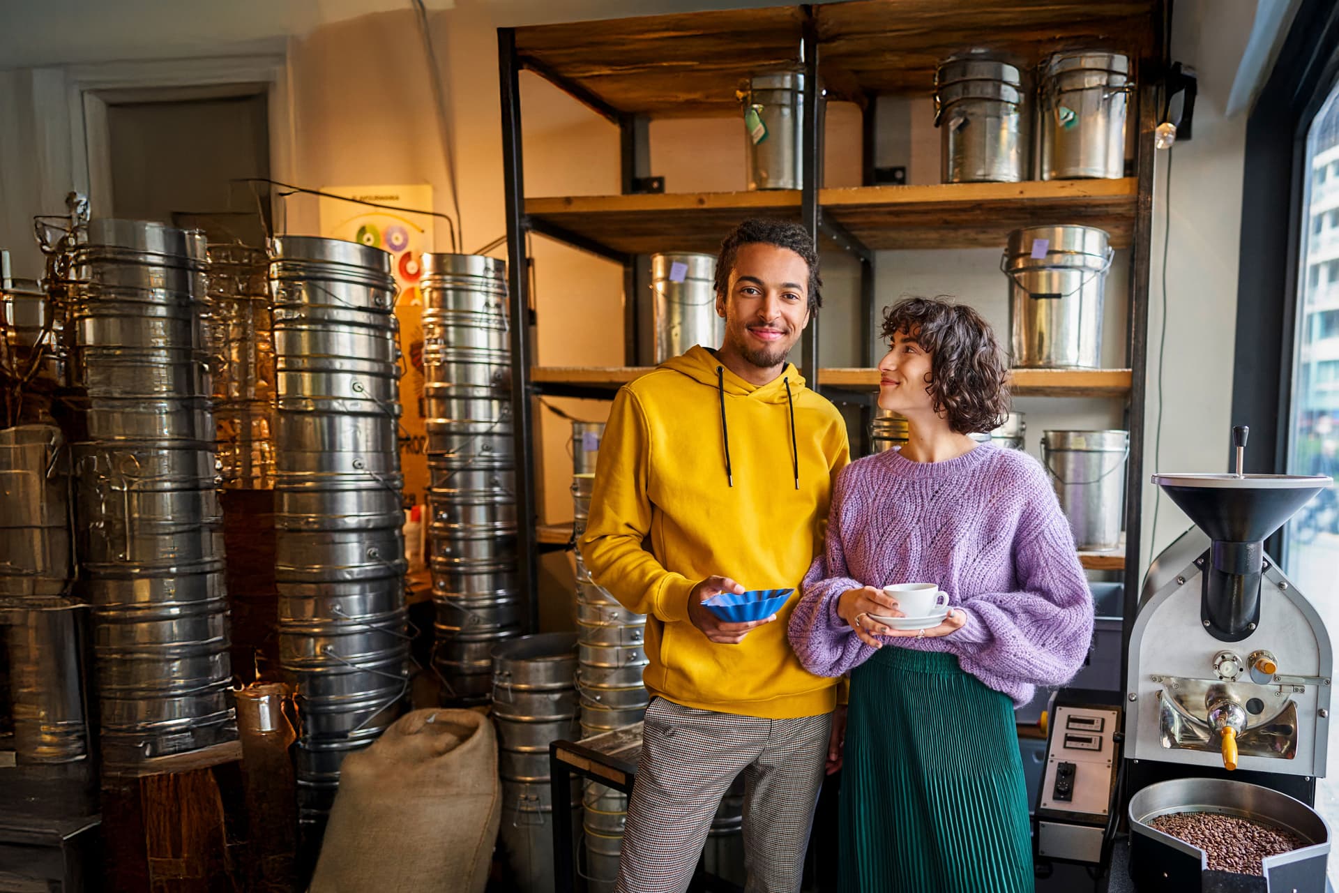Eine junge Frau und ein junger Mann stehen in einer Kaffeerösterei in einem Ladengeschäft. Sie hat eine Tasse in der Hand und er eine Schale mit Kaffeebohnen.