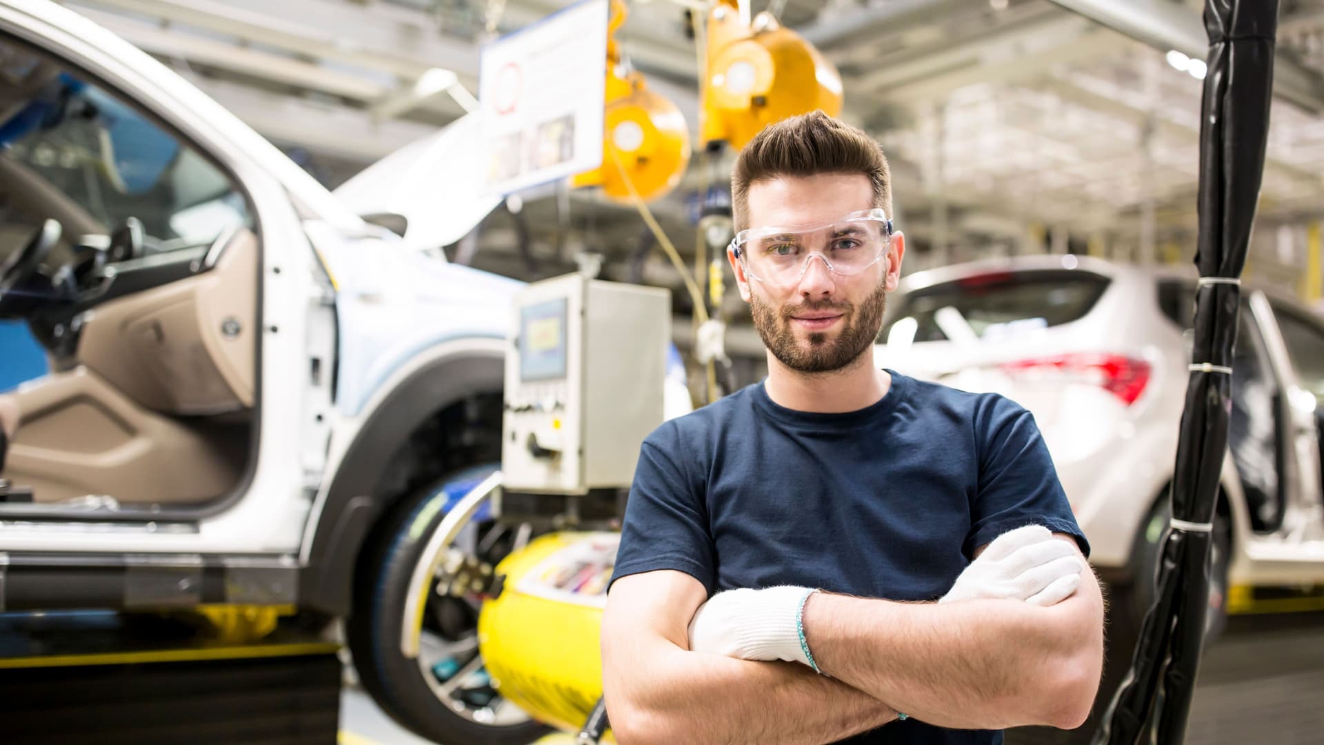 Porträt eines jungen Arbeiters der mit verschränkten Armen und Schutzbrille auf in die Kamera guckt. Er steht in einer Autofabrik.