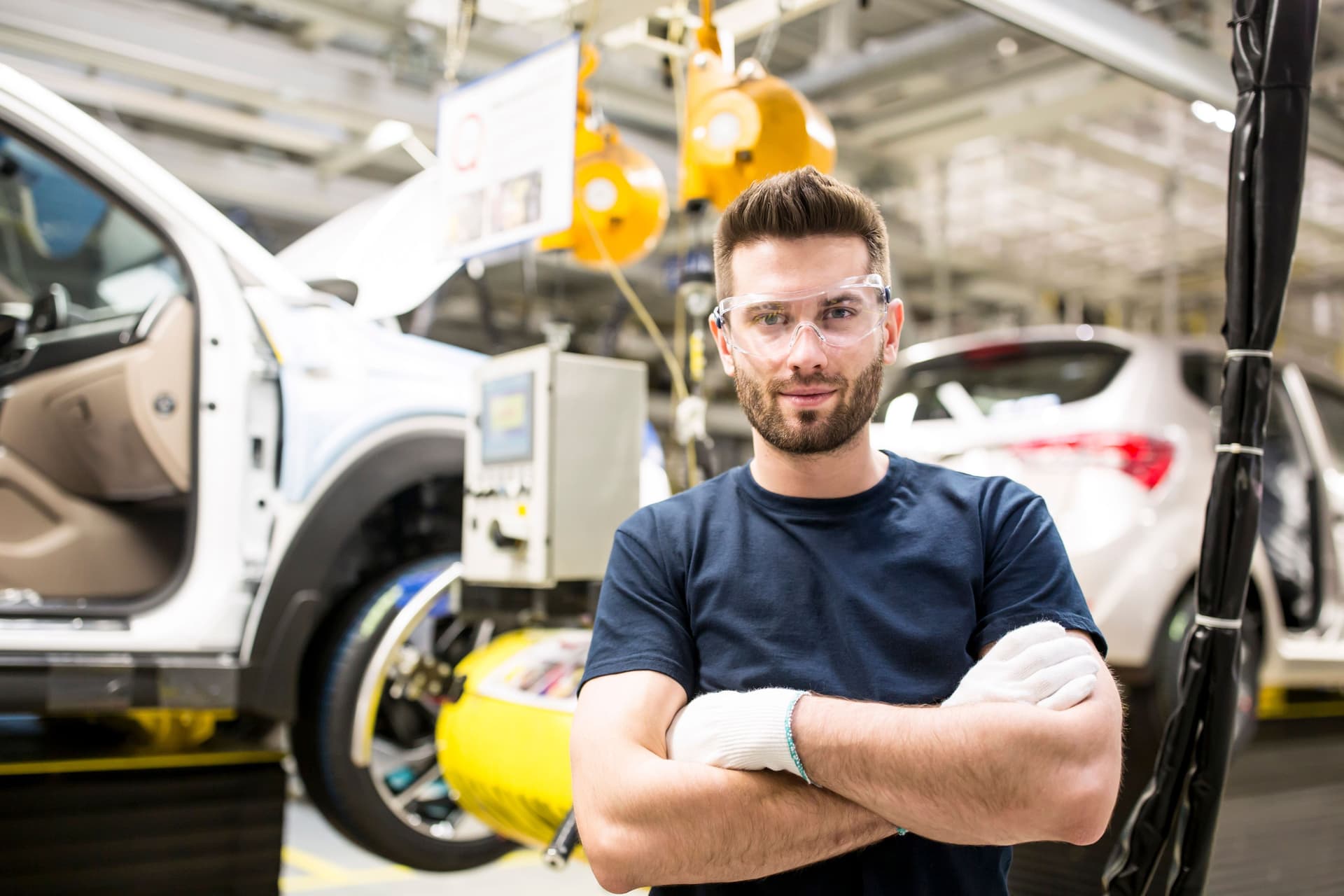 Porträt eines jungen Arbeiters der mit verschränkten Armen und Schutzbrille auf in die Kamera guckt. Er steht in einer Autofabrik.