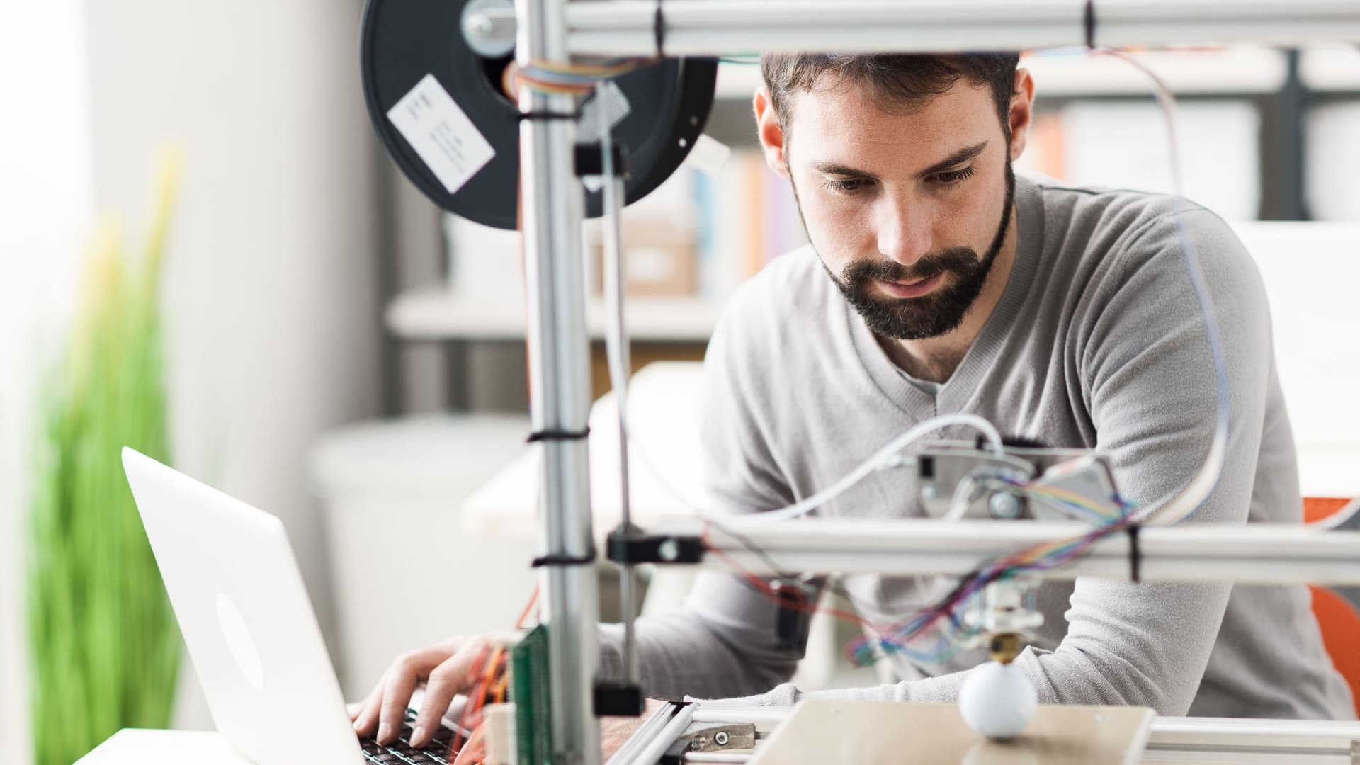 Junger Mann arbeitet an einem Laptop in einem Labor. Er hat einen Prototypen und 3D-Drucker vor sich.
