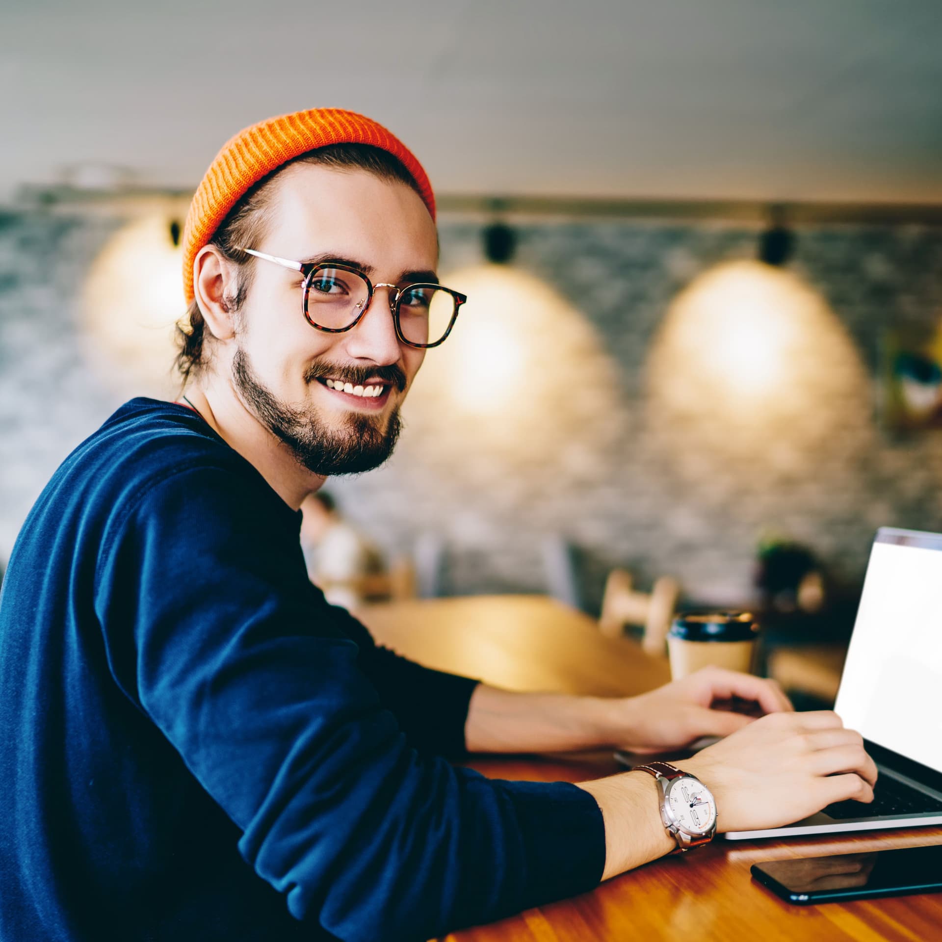 Junger Mann mit Brille und Mütze guckt lächelnd in die Kamera. Er ist in einem Cafe und auf dem Tisch vor ihm ist ein Laptop und ein Smartphone.